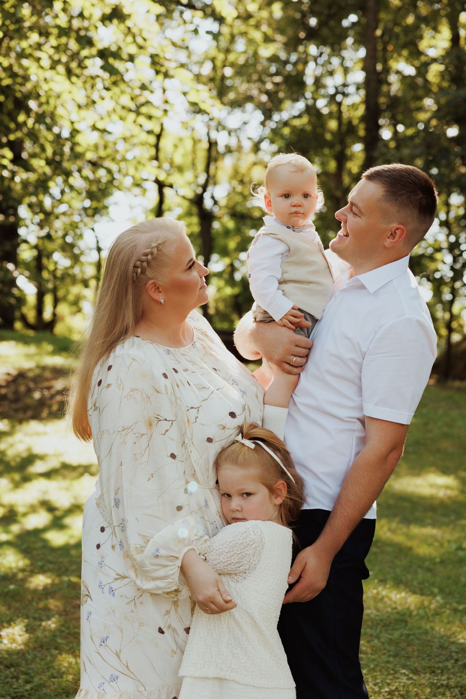 Family wearing coordinated white outfits poses together for outdoor portrait session in sunlit wooded setting.