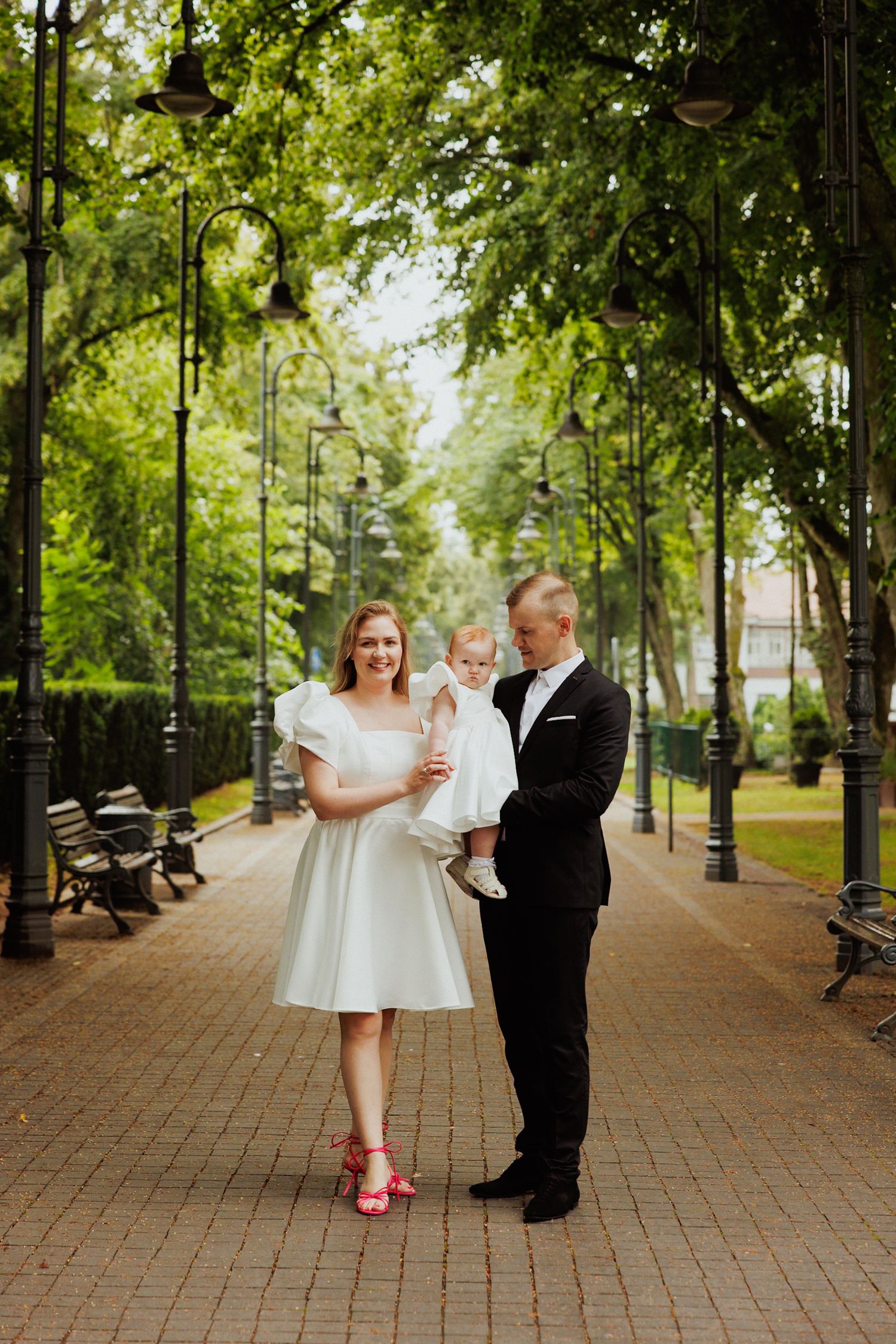 A couple stands on a tree-lined brick path in a park, with vintage lampposts and benches visible in the background.