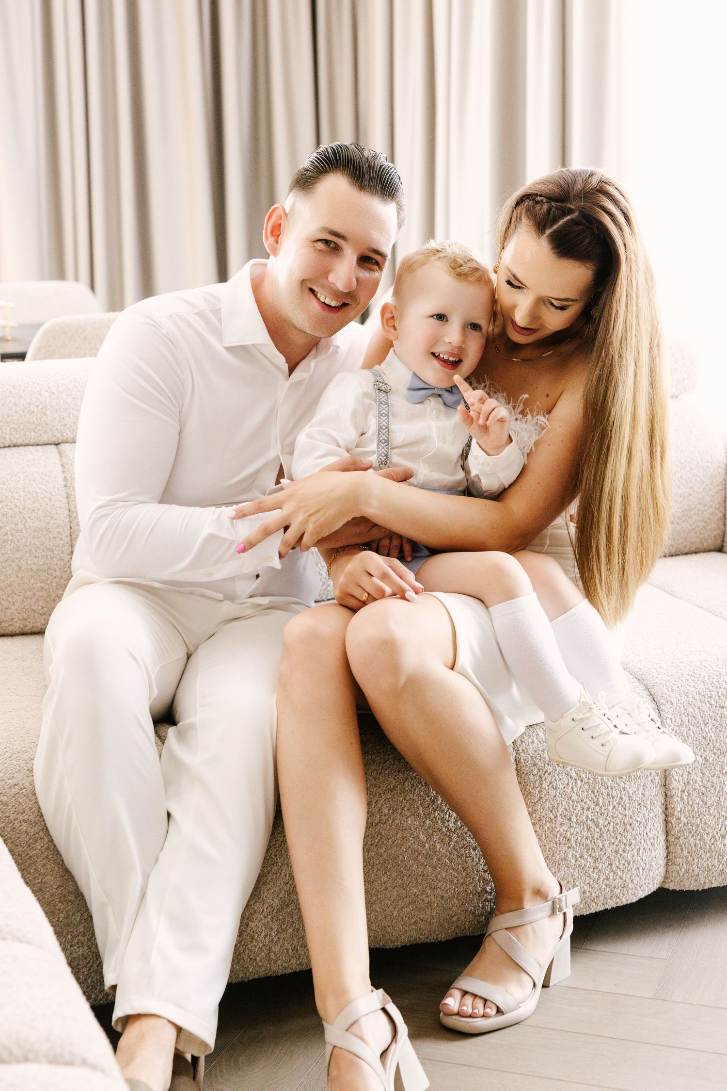 Family in white outfits sharing tender moments together on a light colored couch in bright interior setting.