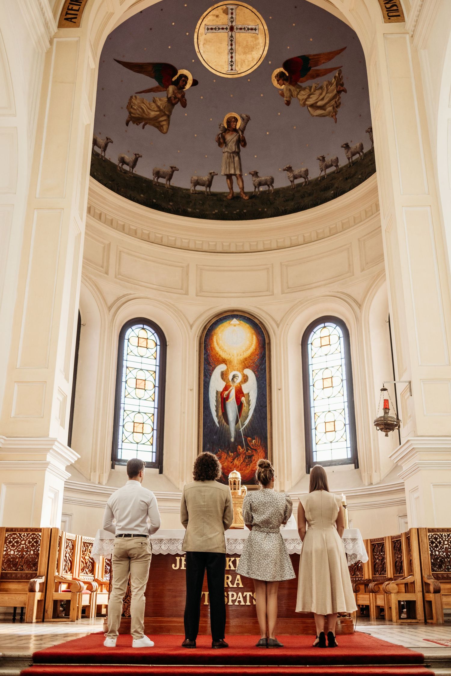 People stand in a church sanctuary beneath a domed ceiling with religious artwork and stained glass windows.