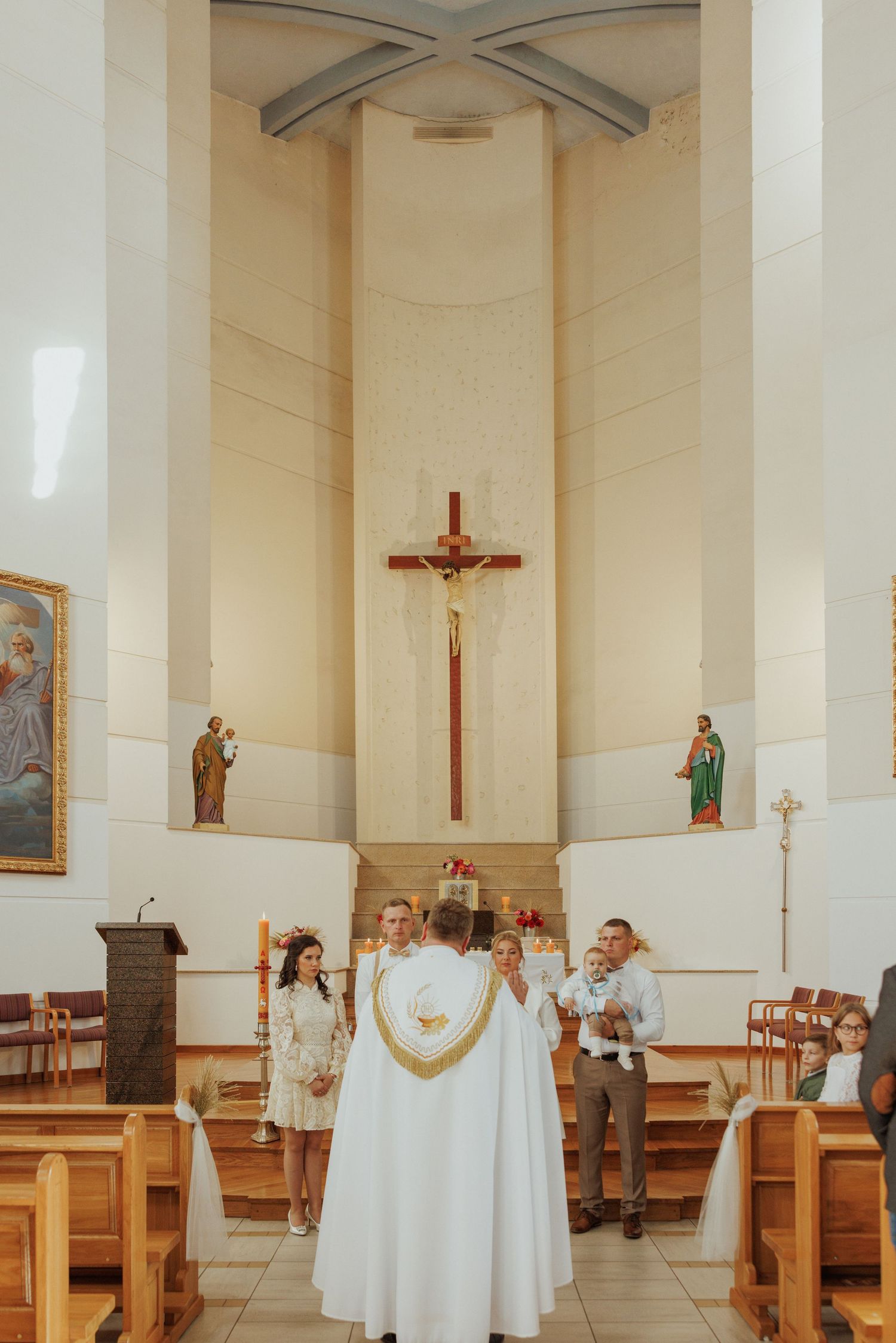 A religious ceremony taking place in a modern church sanctuary with a large crucifix mounted on the cream-colored wall.