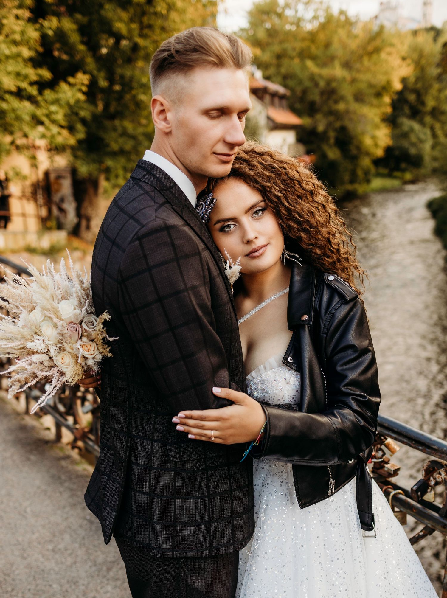 A couple in wedding attire poses by a river, with the bride wearing a white dress and leather jacket holding dried flowers.