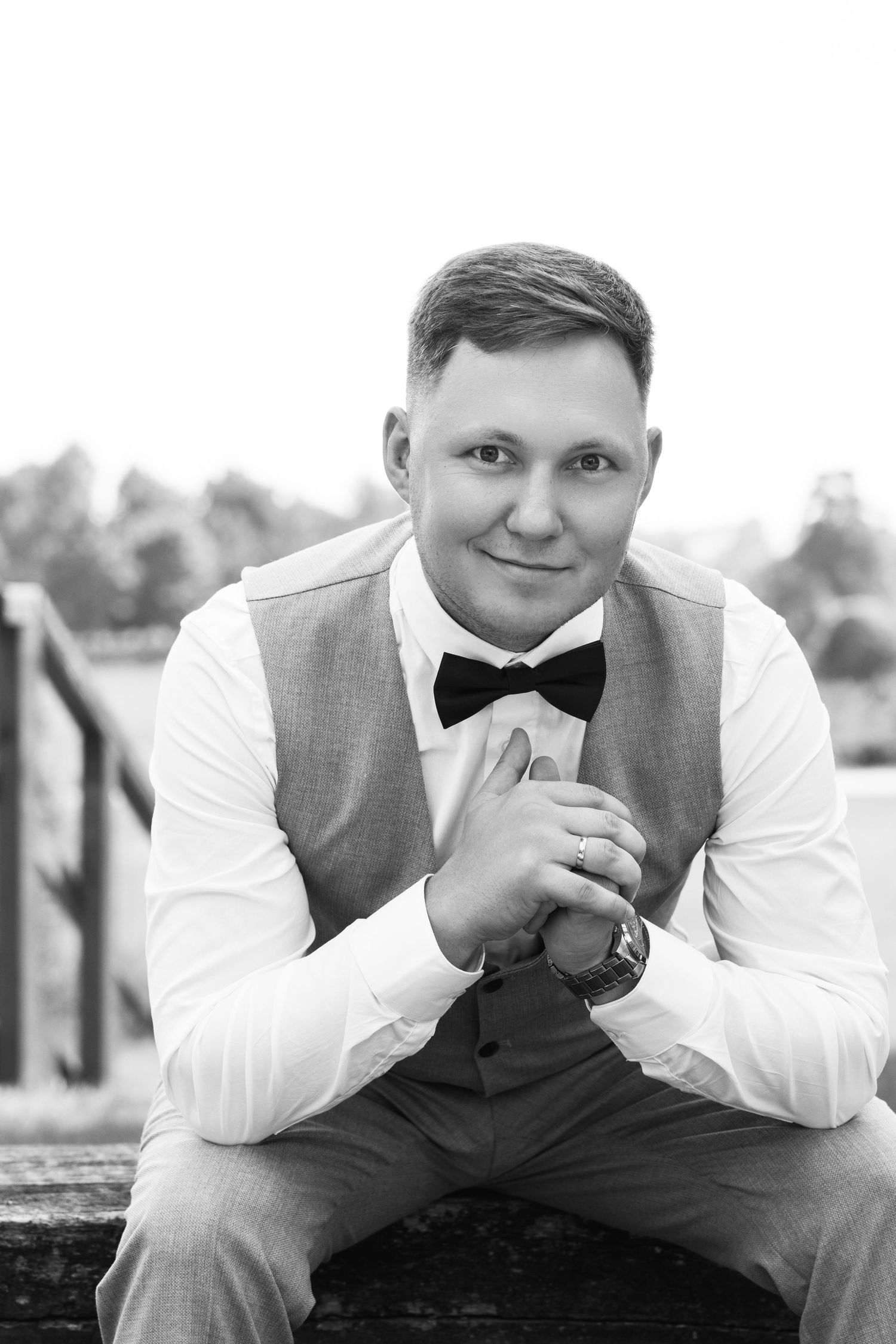 Black and white portrait of a smiling man in a bow tie, vest, and dress shirt, seated outdoors.