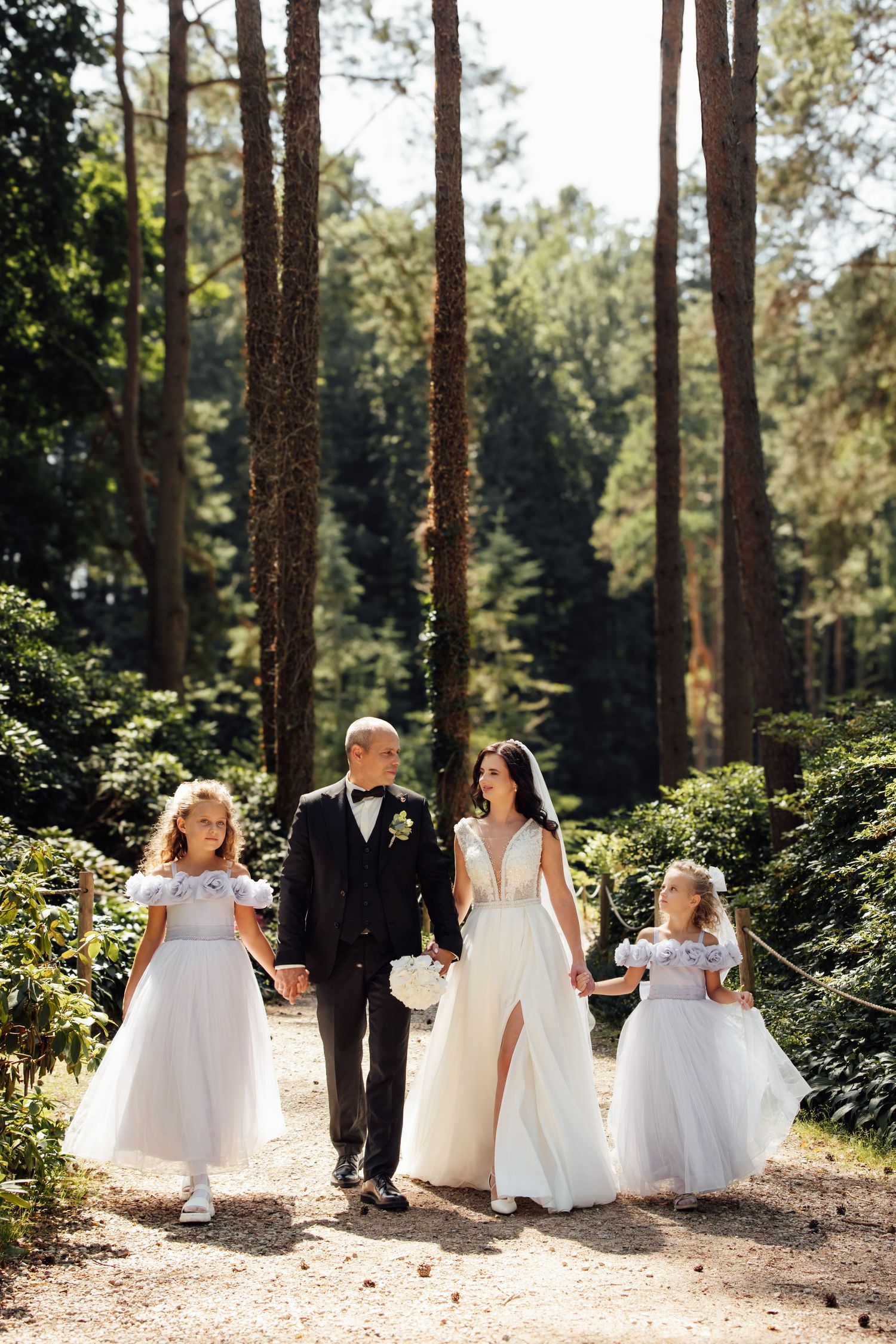 Wedding couple walks with two flower girls in white dresses through a sunlit forest path.