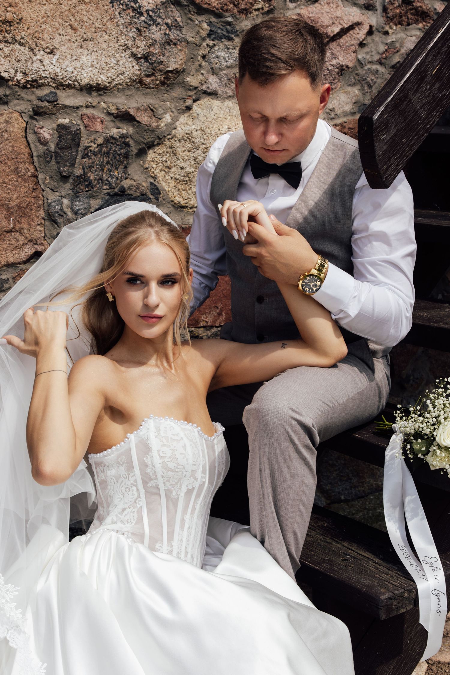 Elegant bride in white strapless gown and veil sits on stone steps as groom in gray vest kisses her hand.