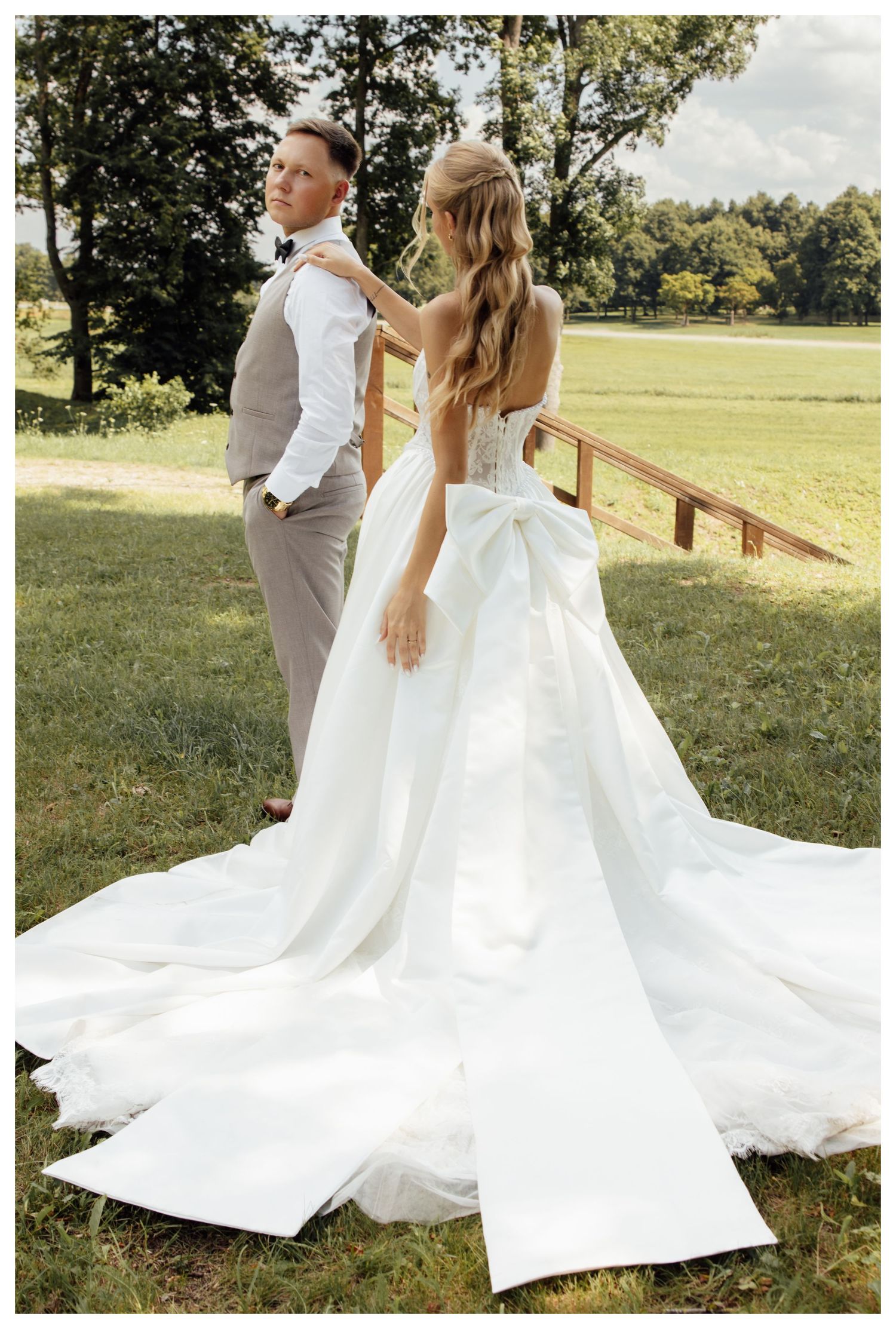 Bride in white gown with large bow adjusts grooms gray suit jacket outdoors near wooden fence.