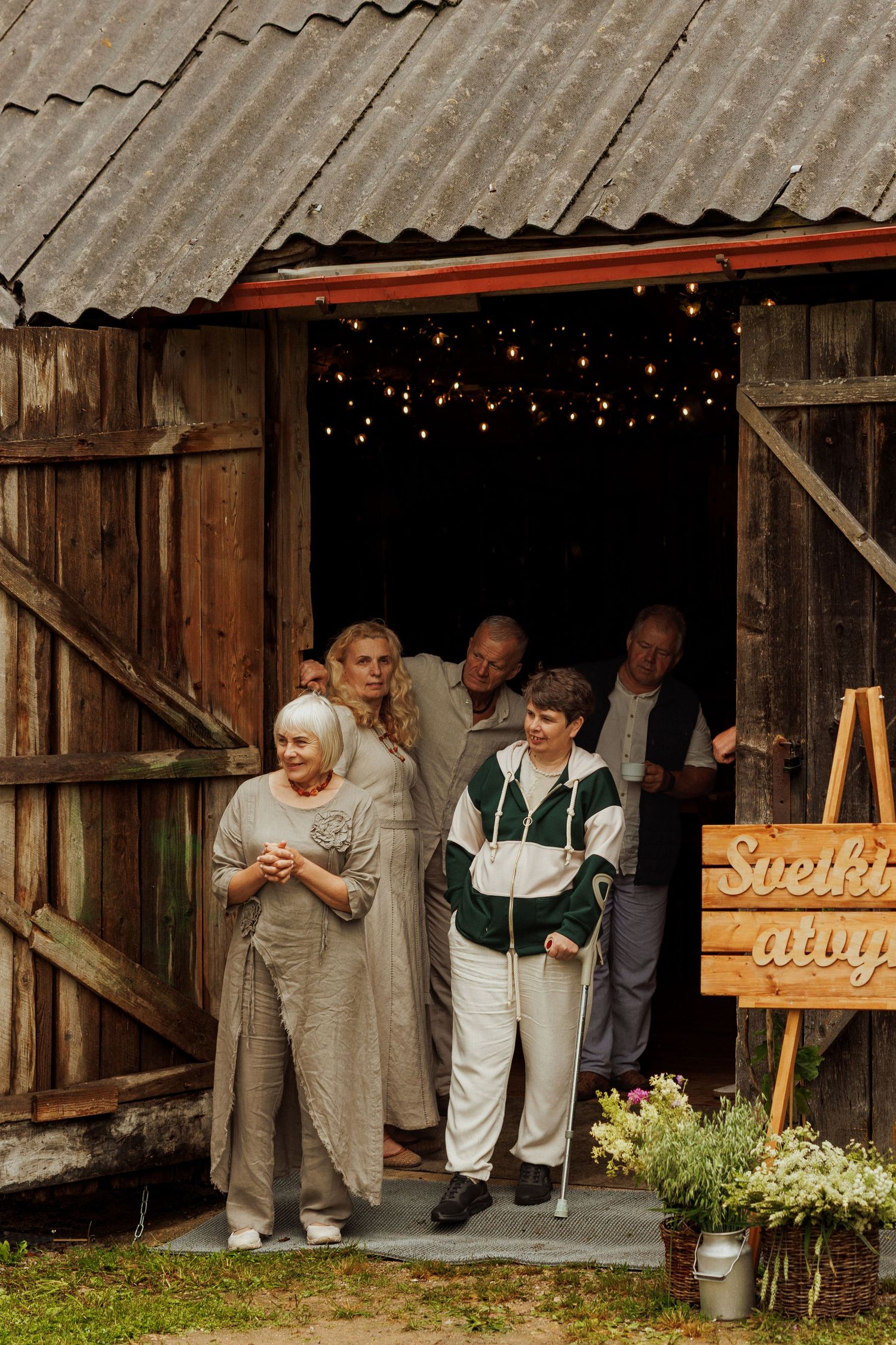 Group of elderly people emerging from a rustic wooden barn decorated with string lights and a welcome sign.