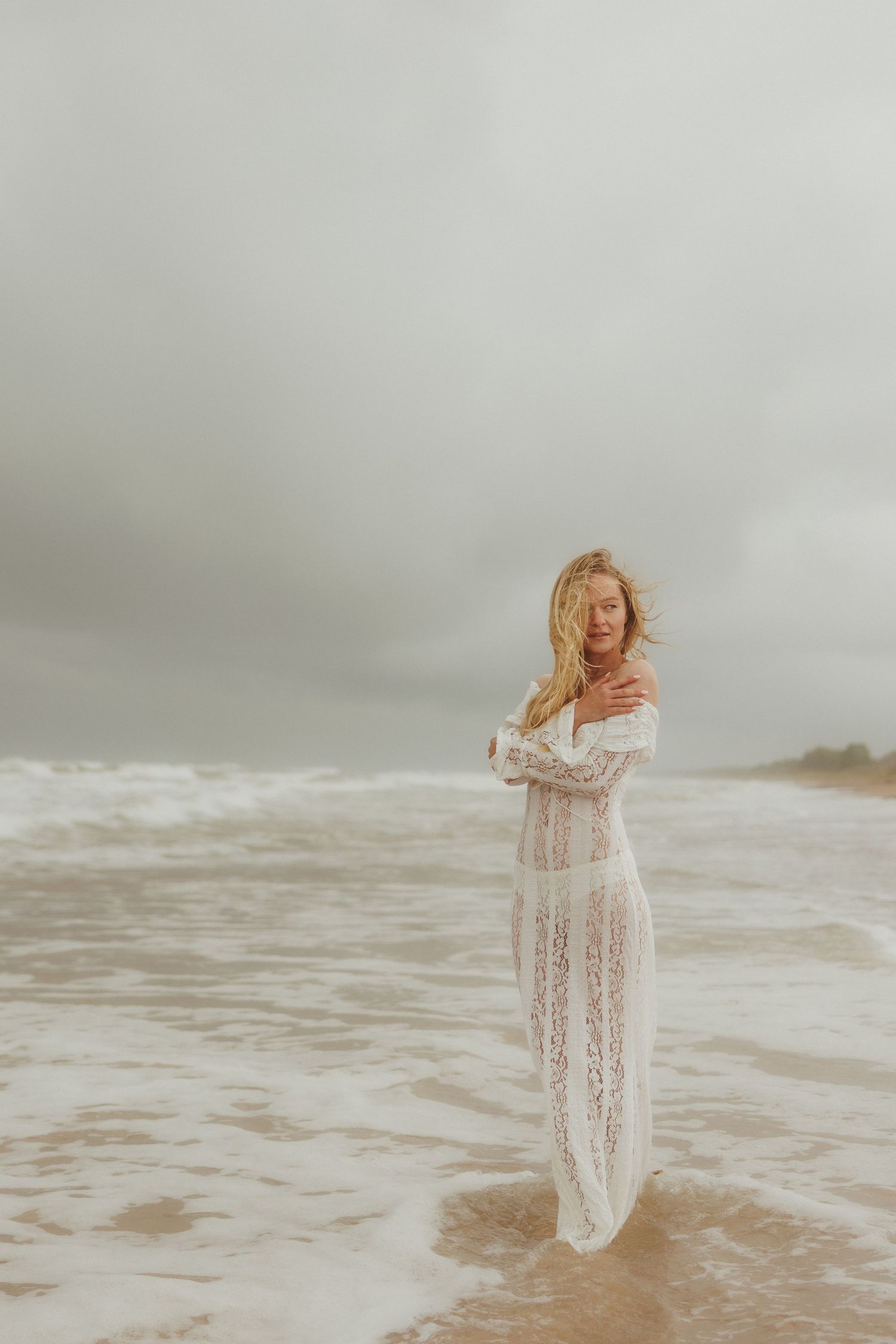 Blonde woman in sheer white lace gown standing in ocean waves on overcast beach.