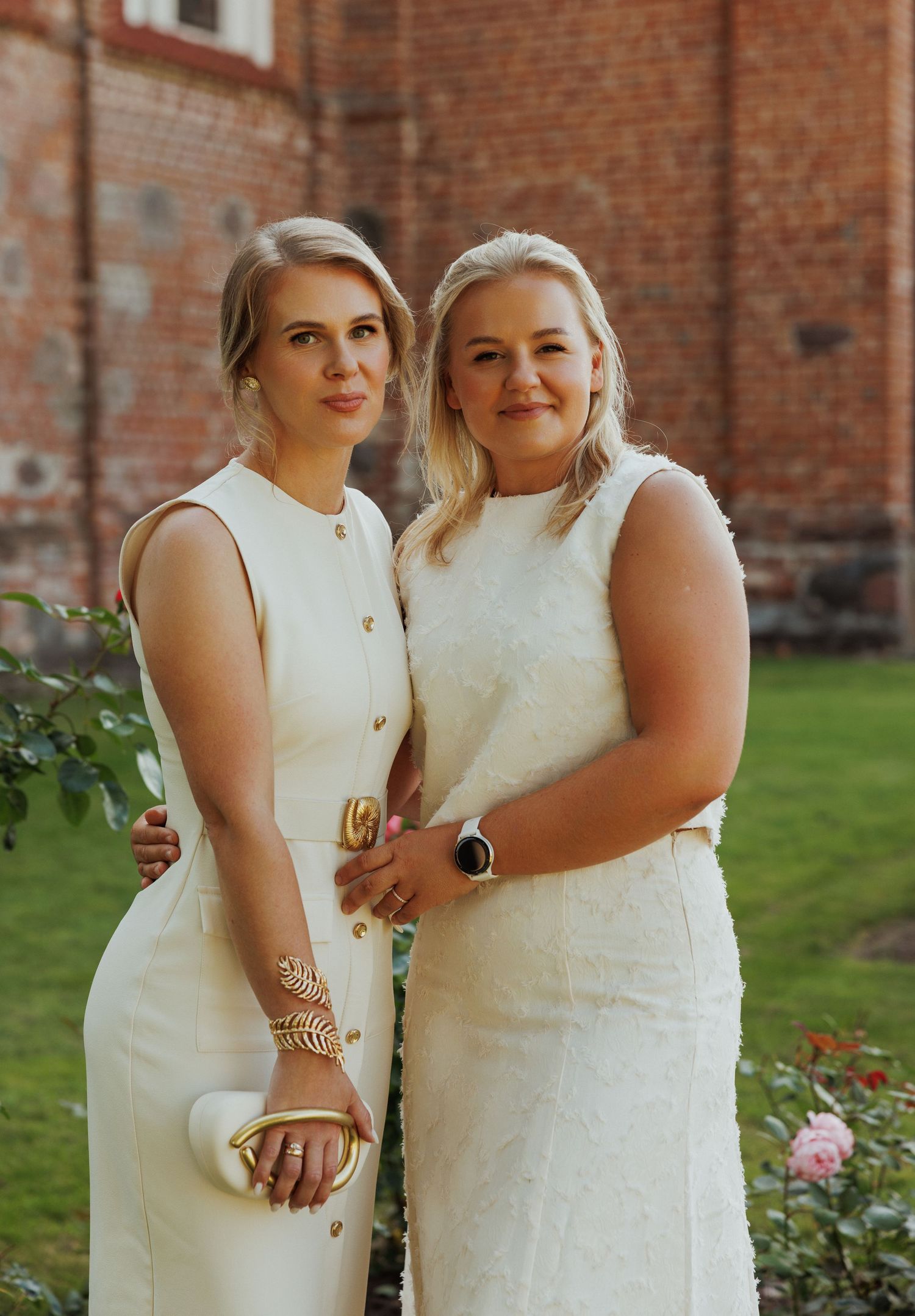Two people in white summer outfits stand close together in front of a brick building with roses.