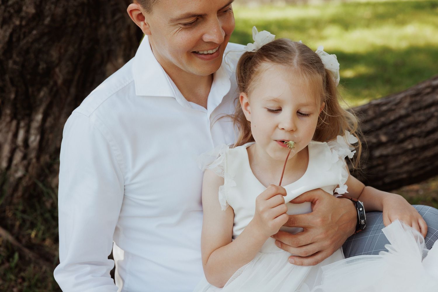A parent and child share a tender moment outdoors while sitting on a fallen tree trunk in a sunlit garden setting.