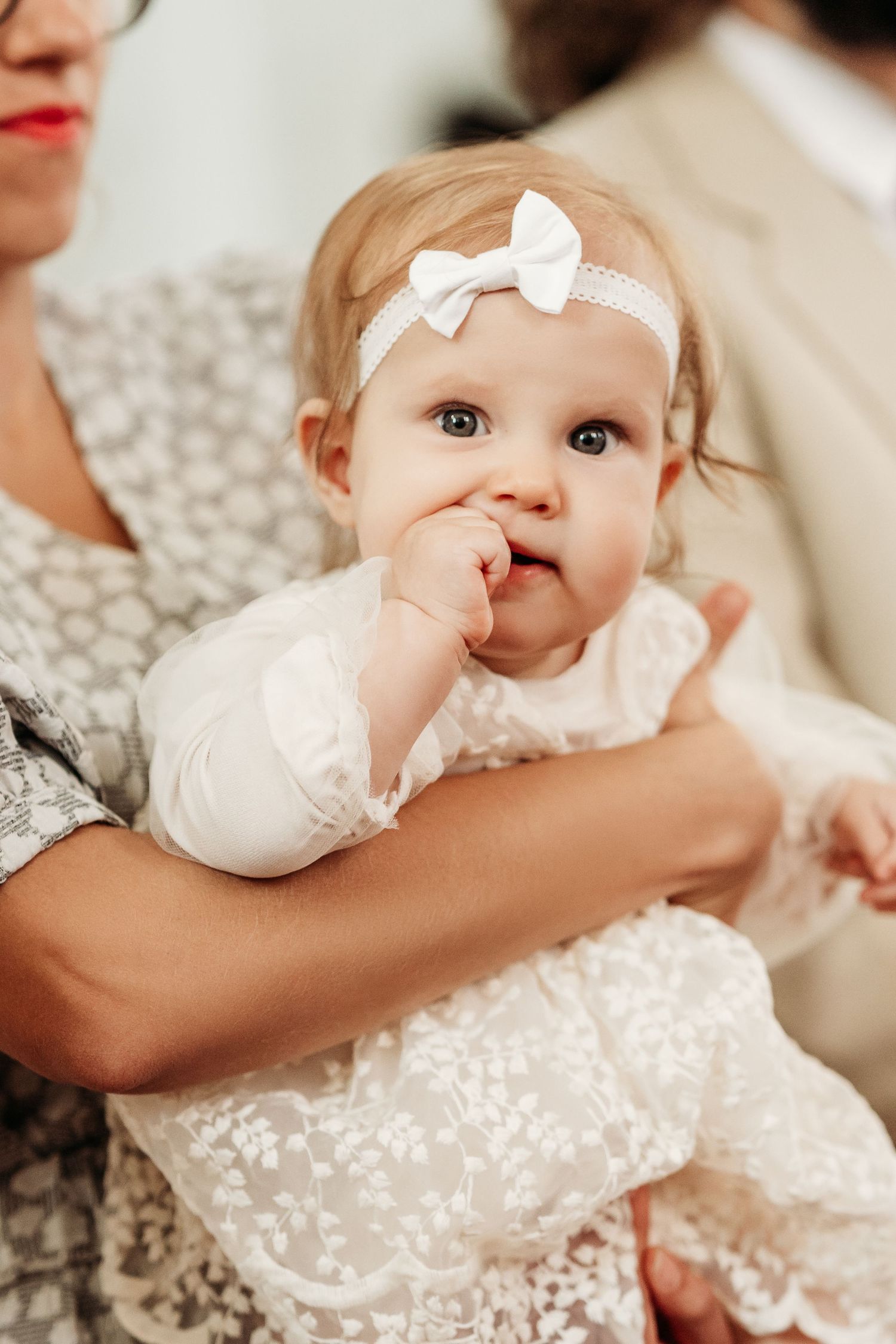 Baby in white lace dress with bow headband being held against light colored background.