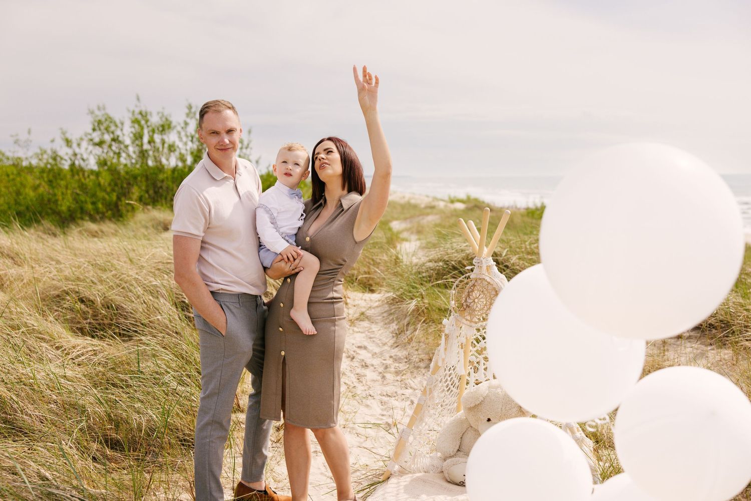 Family walking together on a sandy beach path with white balloons against a natural coastal landscape.