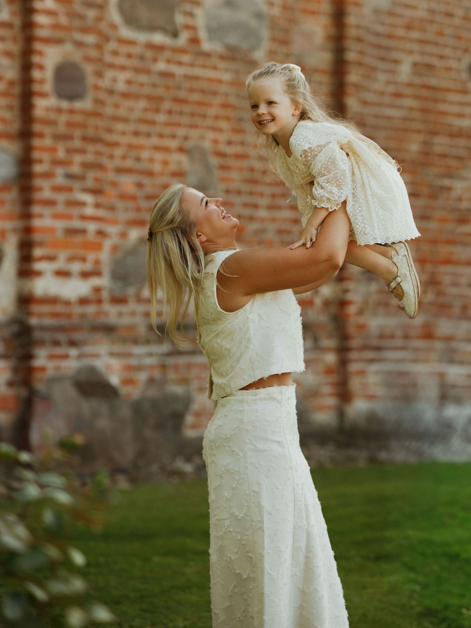A woman in a white dress lifts a small child in the air against a rustic brick wall background.