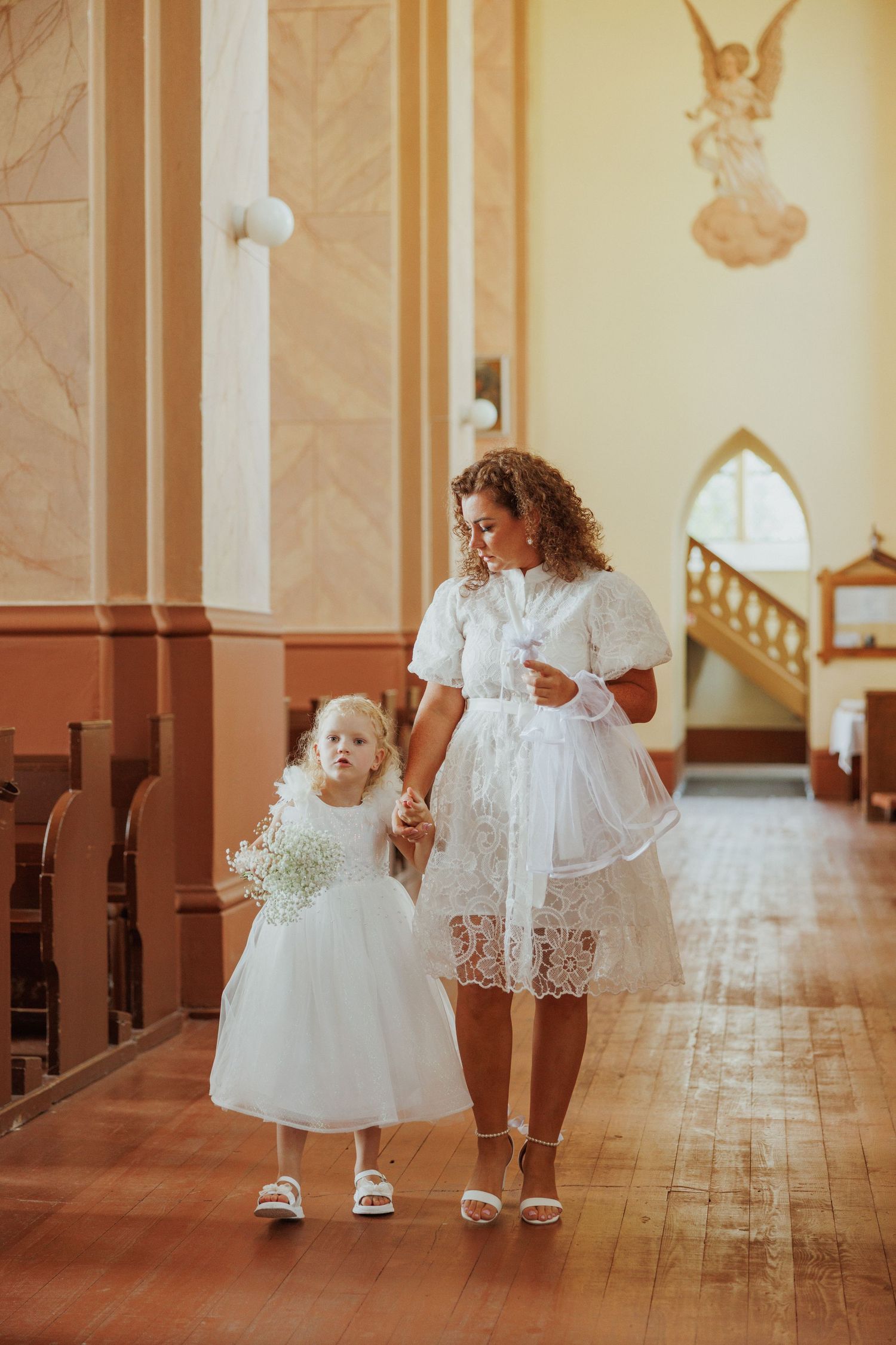 Two children in white christening gowns walk down a church aisle with wooden pews and ornate decor.