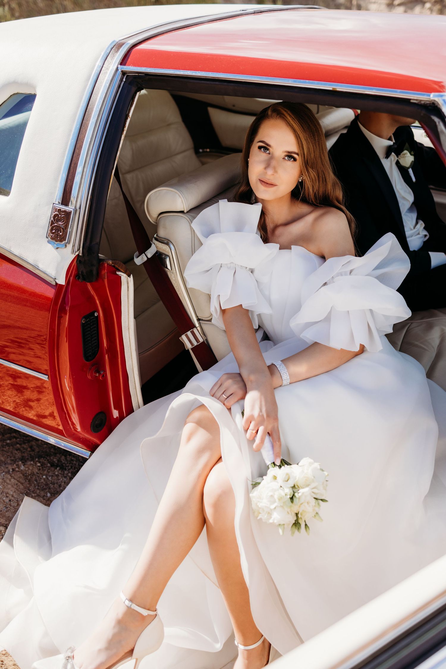 Bride in white wedding dress poses elegantly inside a vintage red car showing different angles.