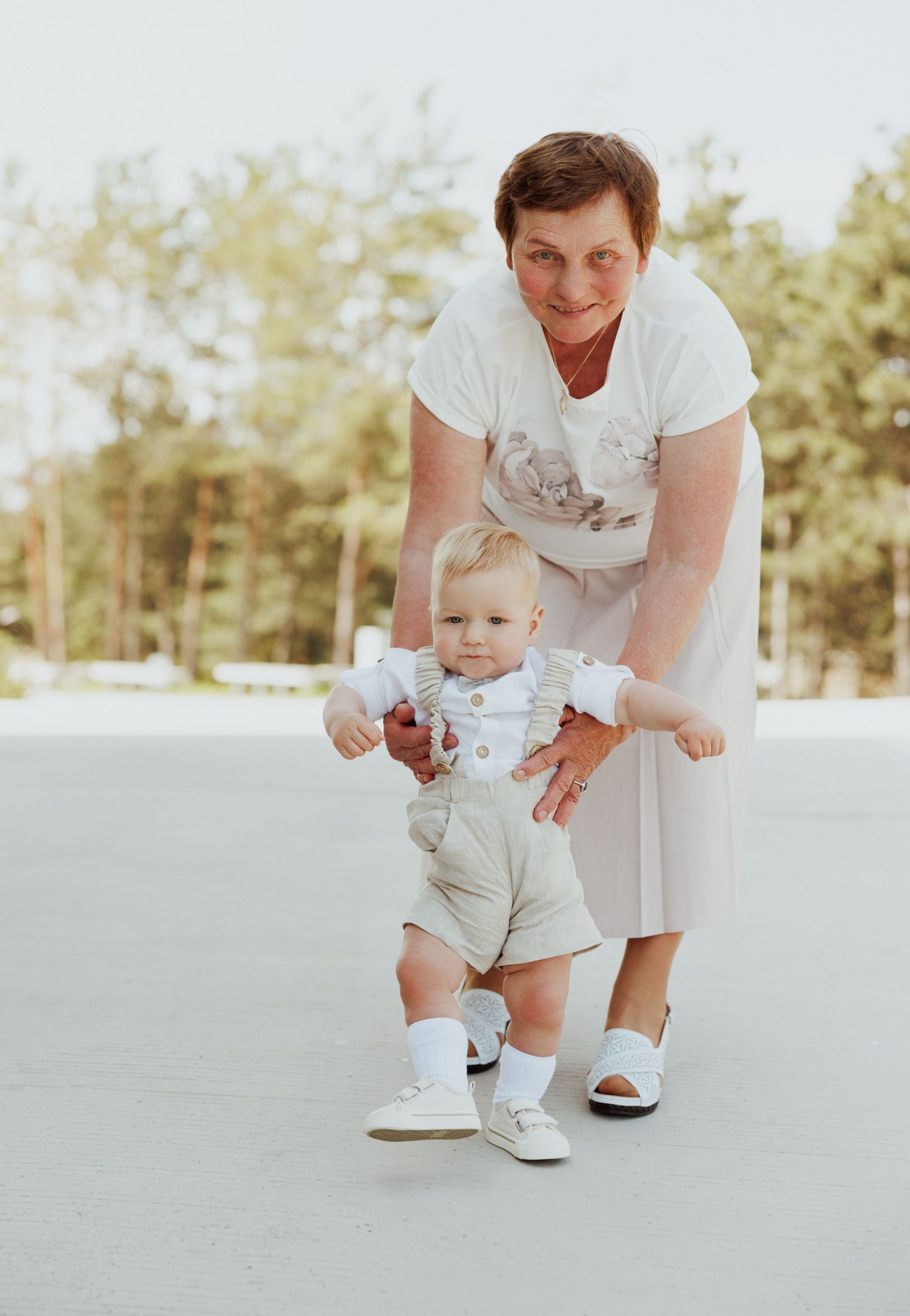 Baby in light colored outfit learning to take first steps with support on bright outdoor surface.