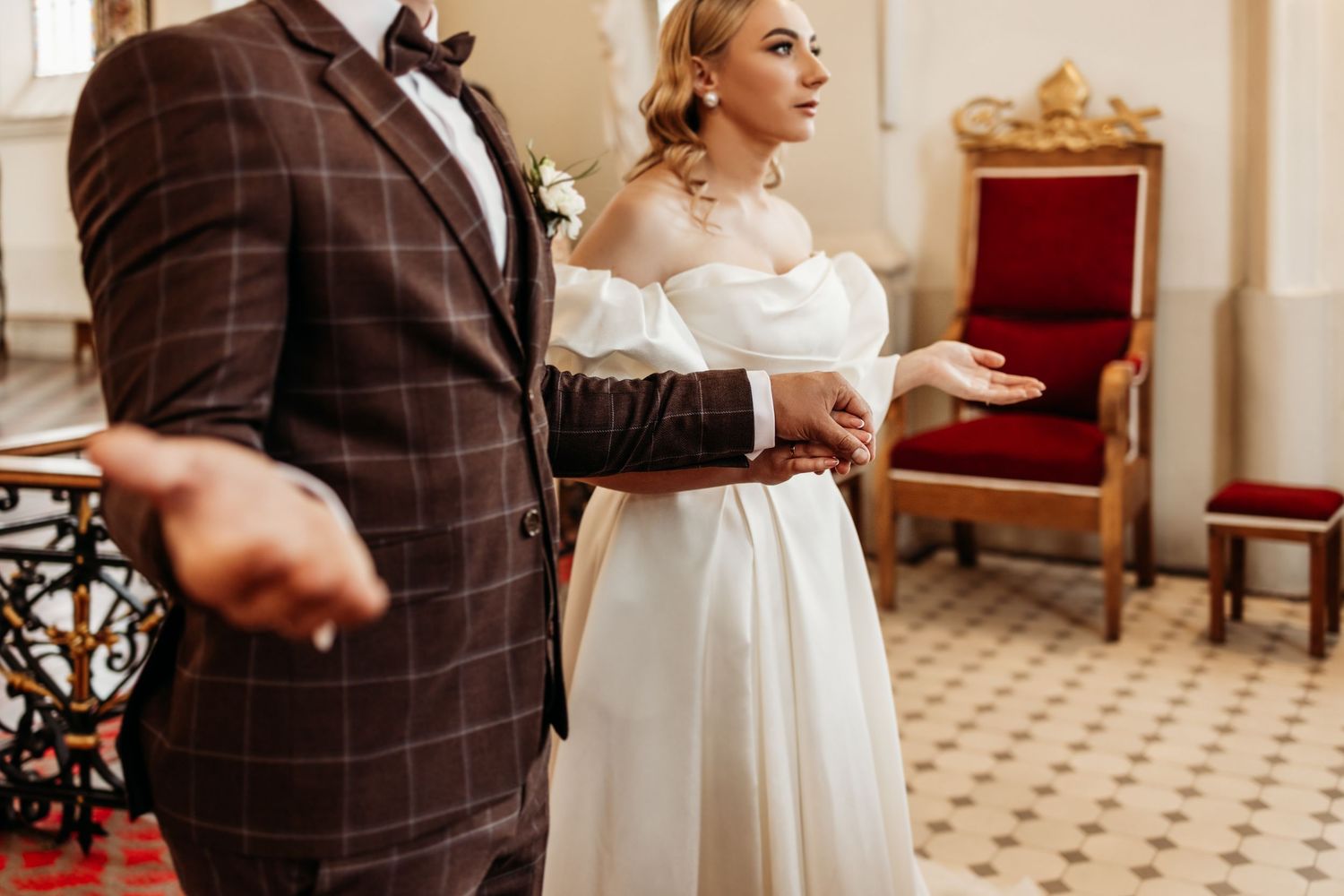 A couple in wedding attire stands in an elegant room with a red velvet chair and tiled floor in the background.