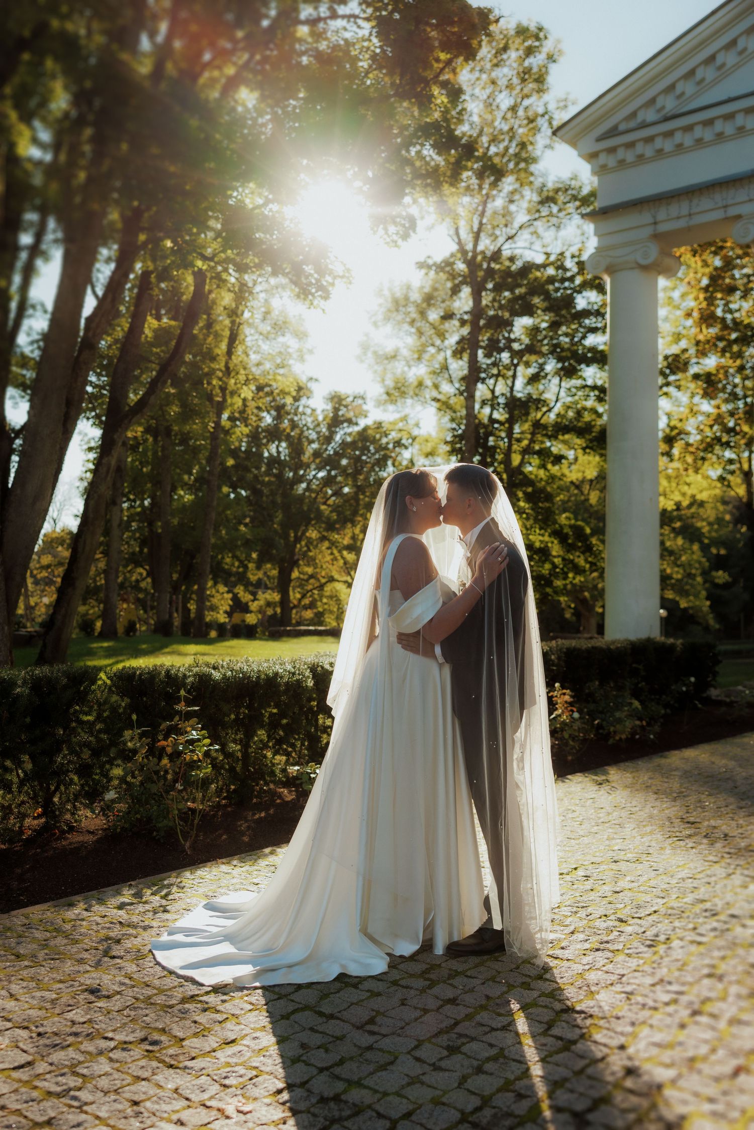 Sunlit wedding portraits against white columns with dramatic flowing veil.