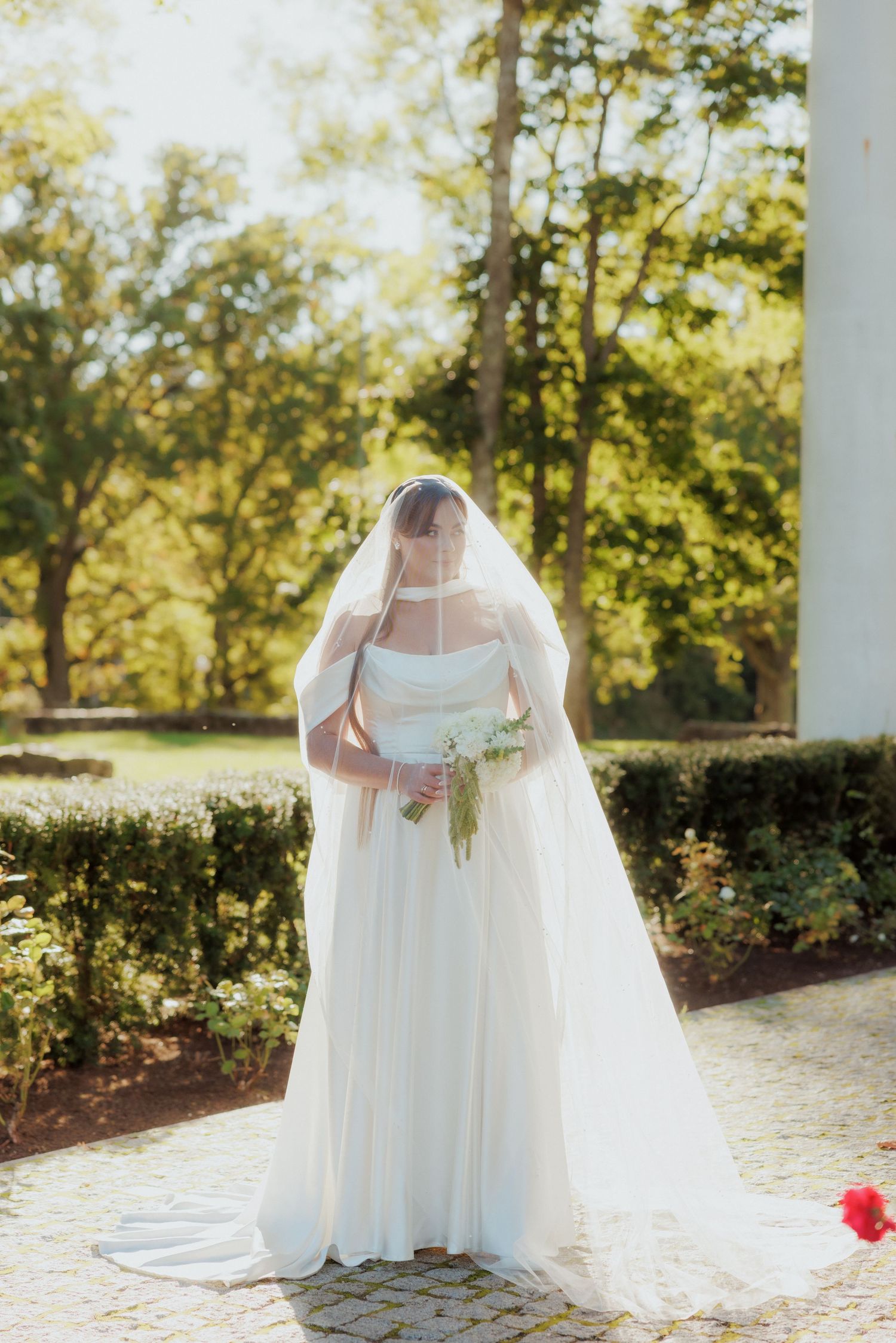 Elegant white wedding dress with flowing veil photographed in a garden setting.