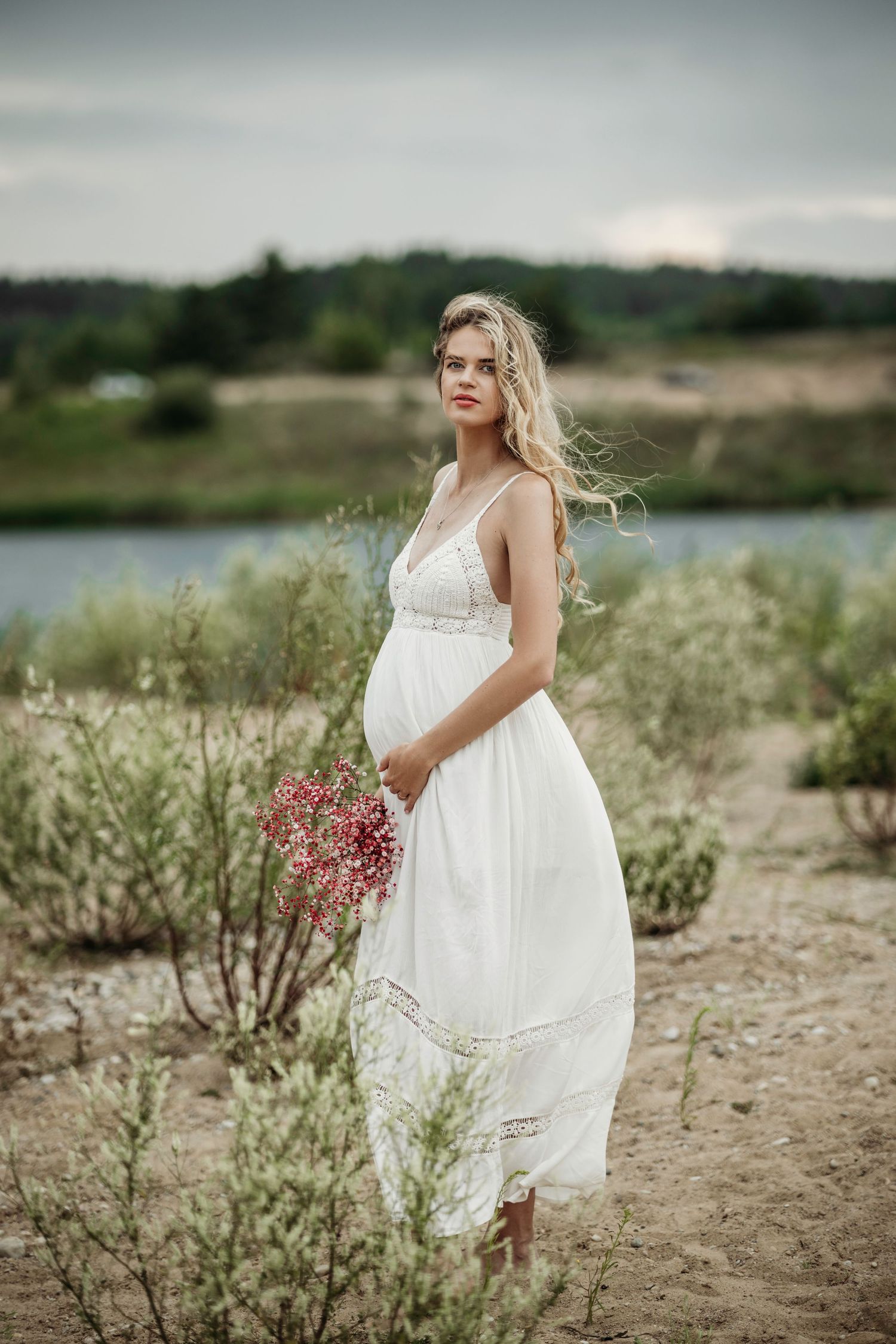 Woman in flowing white dress poses among wildflowers near a lake.
