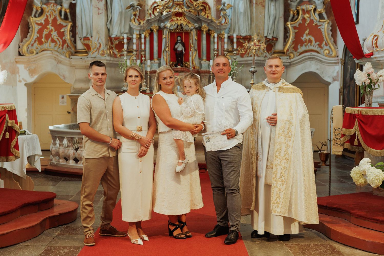 A group stands together in formal attire inside an ornate baroque church with red drapes and gilded altar decorations.