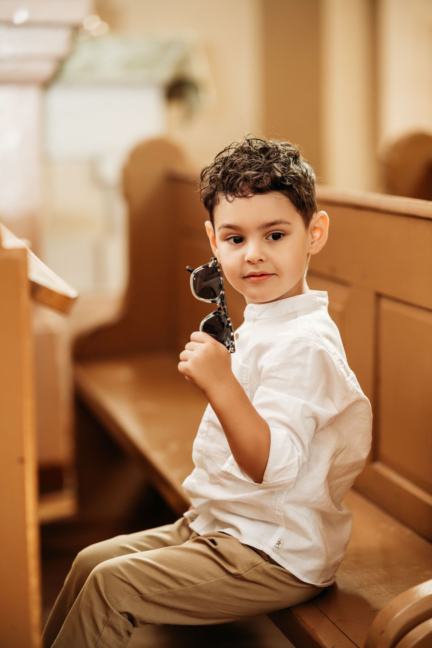 Child in white shirt and khaki pants sitting on wooden church pew holding a small object.