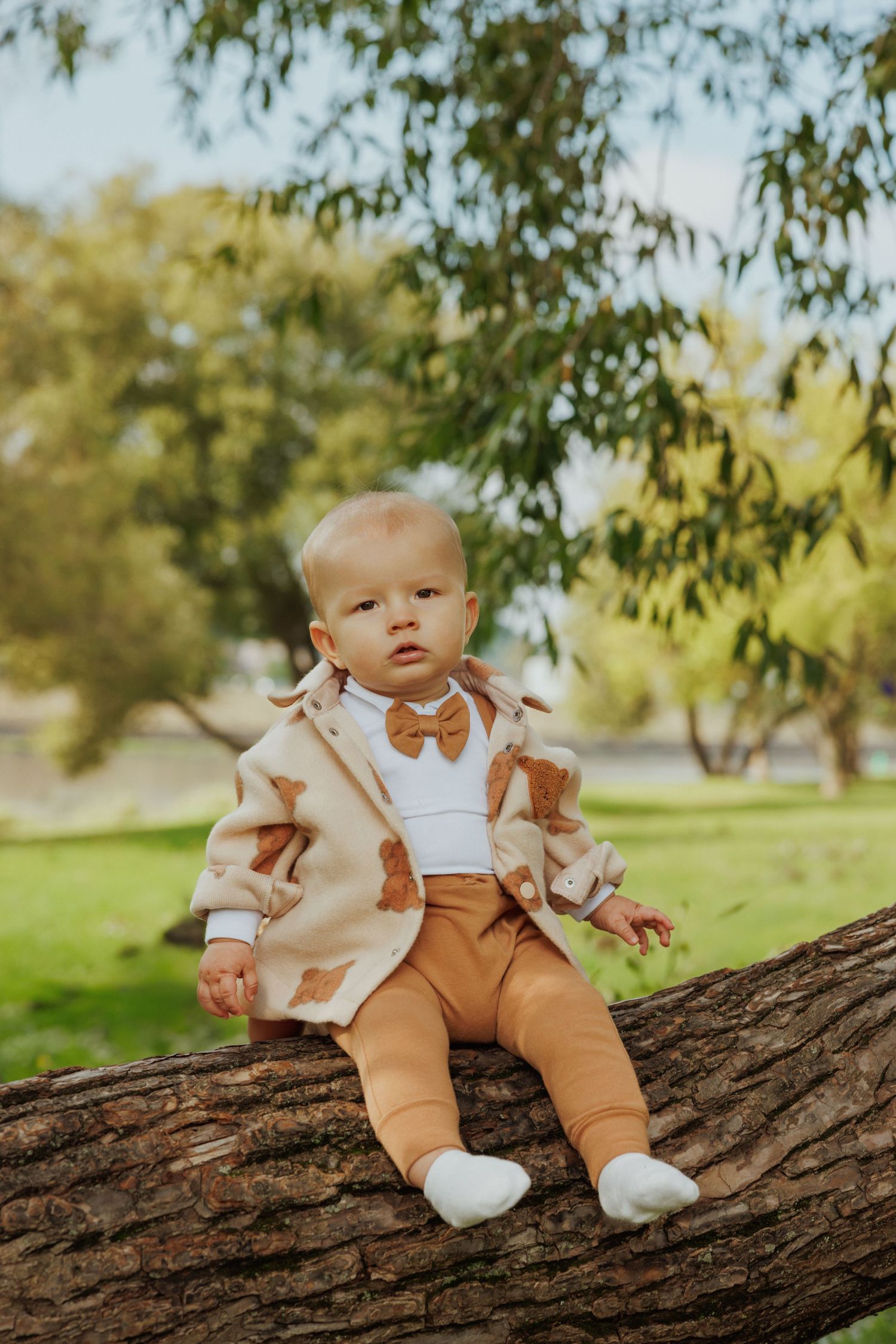 A small baby in brown overalls and white shirt sits on a large tree branch in a sunny outdoor setting.