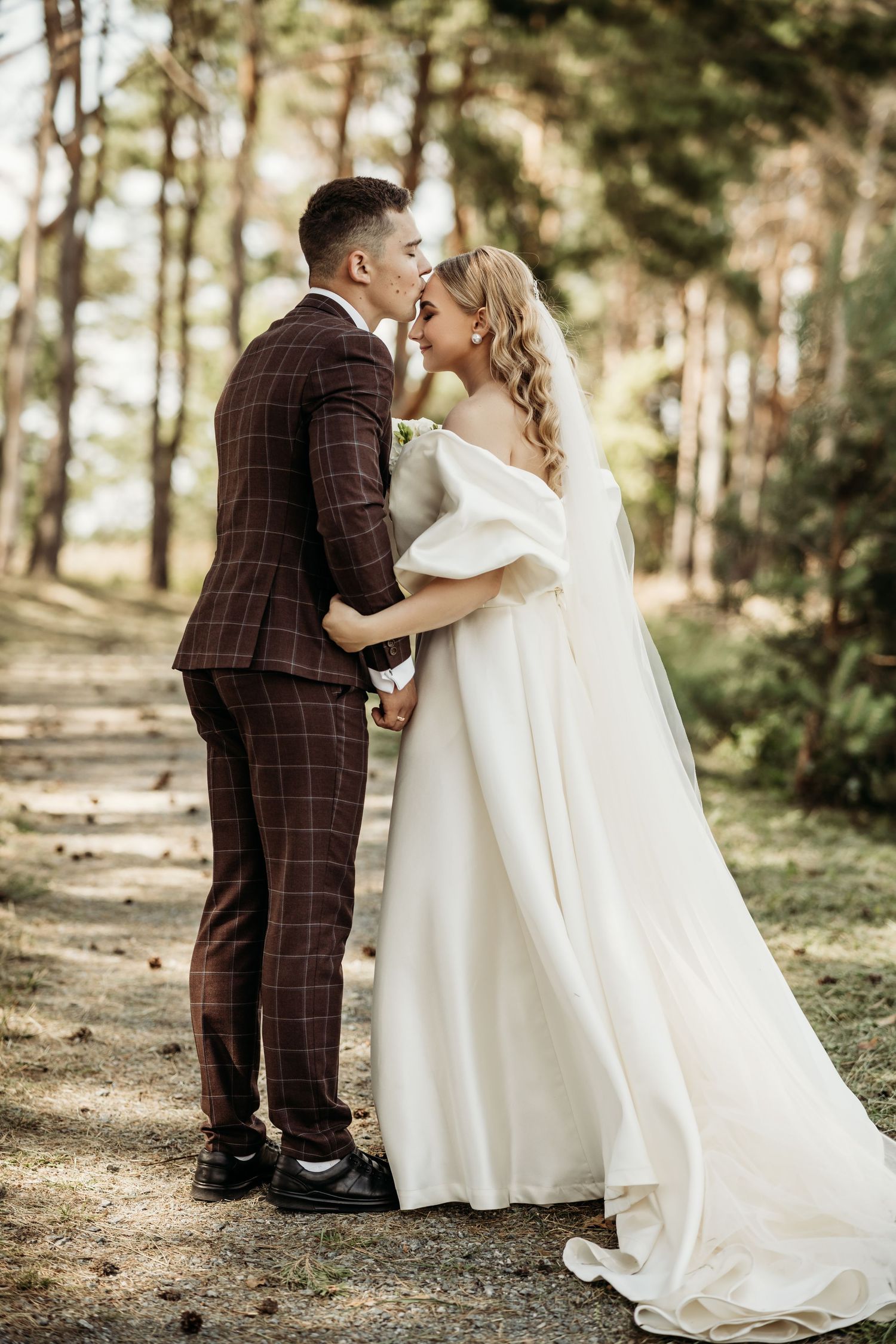 Newlyweds share romantic moments in a sunlit forest setting with pine trees in the background.