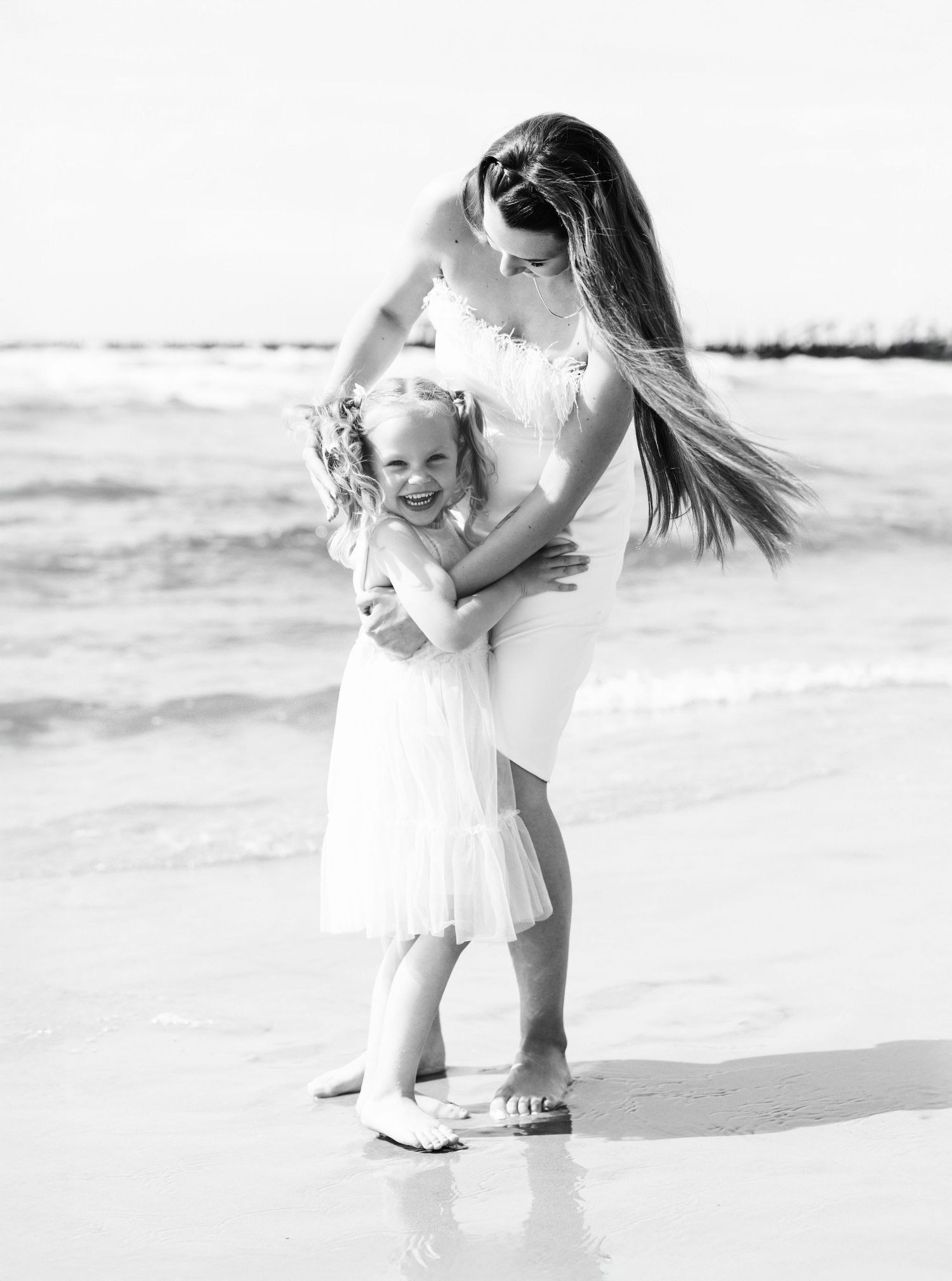 Mother and daughter in white dresses playing together at ocean's edge.