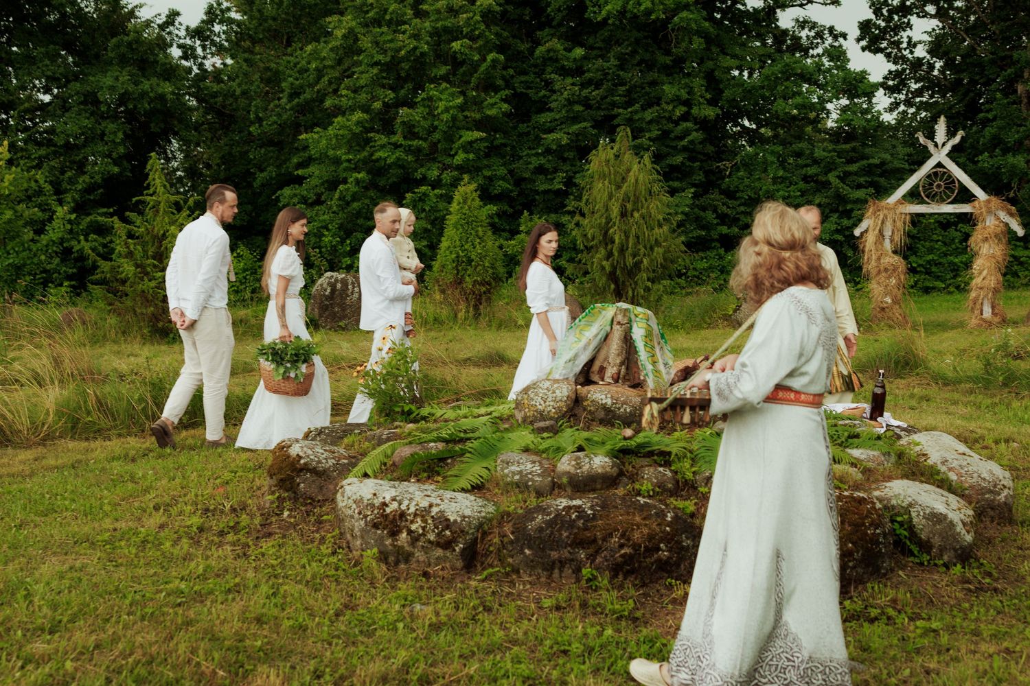Group of people dressed in white performing a traditional outdoor ceremony near stone ruins in a green meadow.