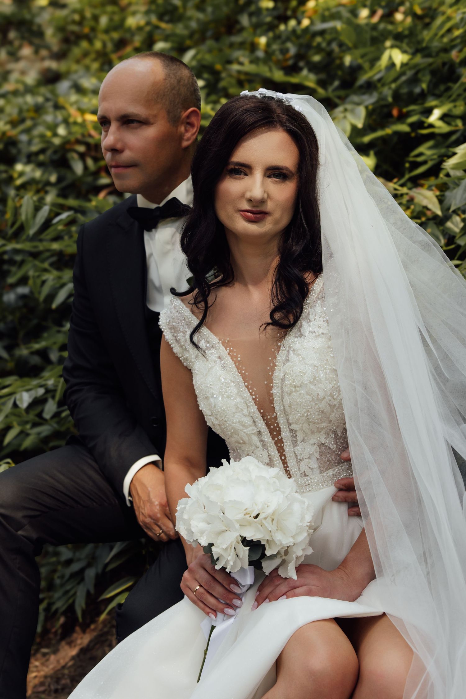 Elegant bride in lace gown with veil holds white bouquet beside groom in black tuxedo outdoors.