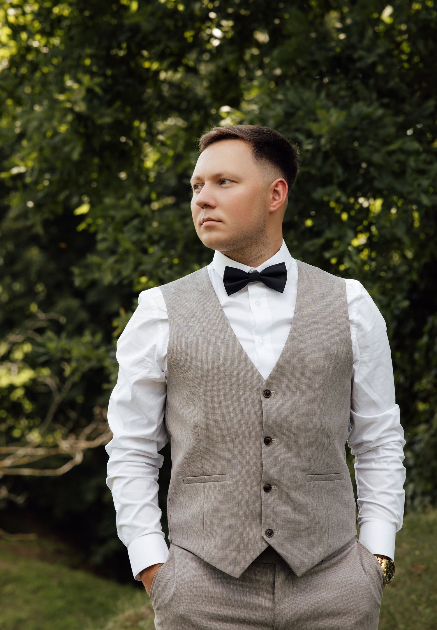 A well-dressed groom in a beige vest, white shirt, and black bow tie poses outdoors among lush green trees.