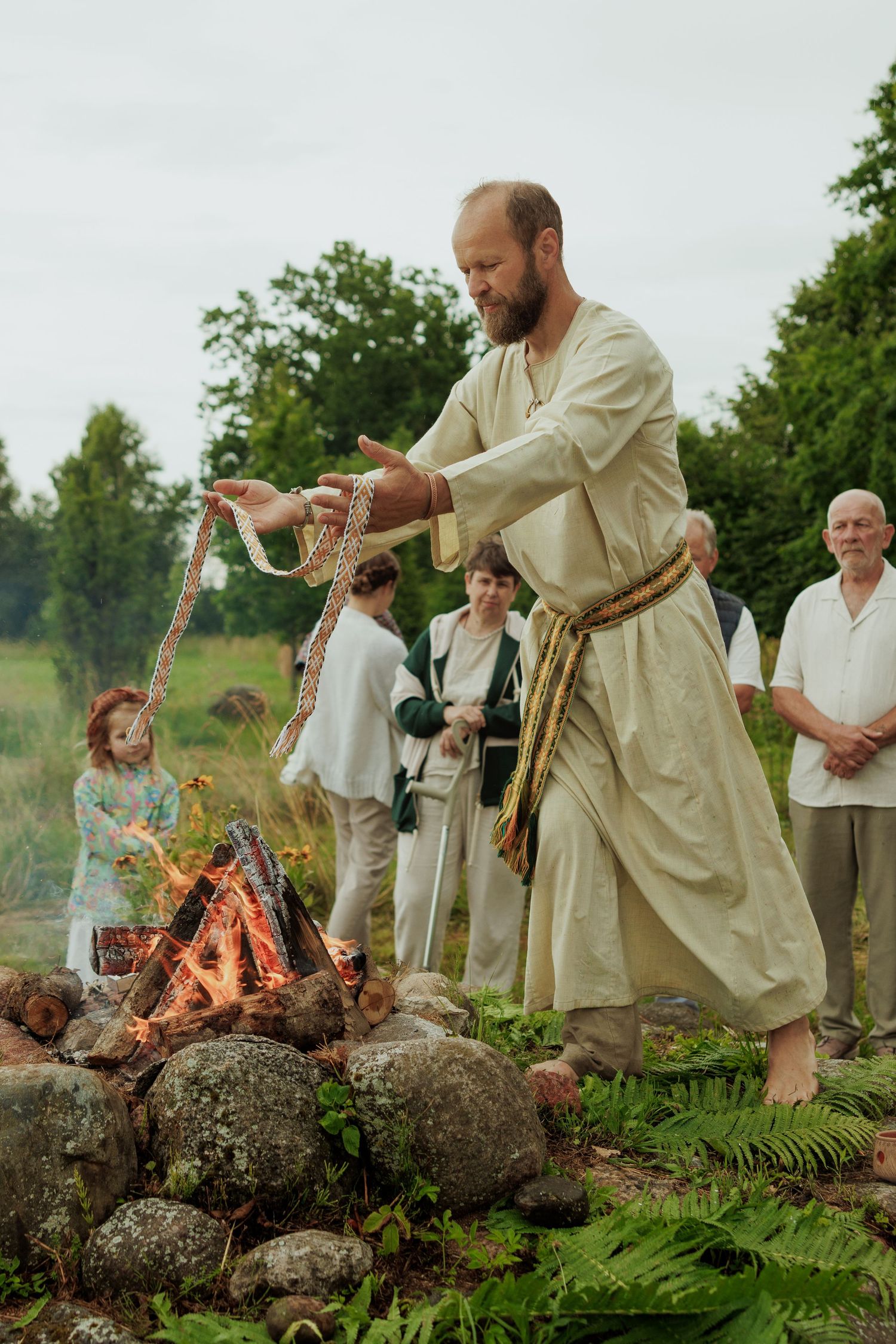 Bearded man in traditional linen robe performing folk ritual with woven belt near stone campfire.
