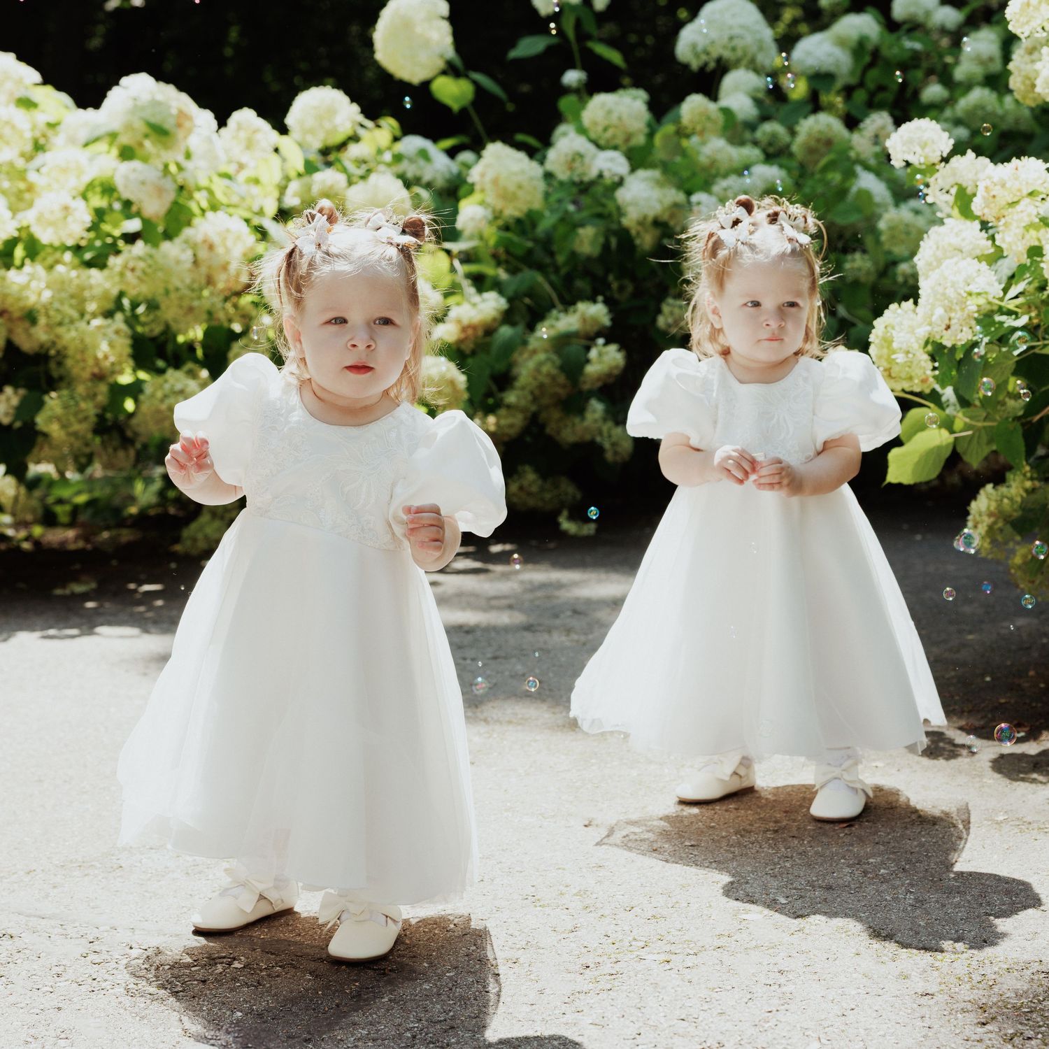 A row of flower girls in white dresses stand among blooming hydrangea bushes in a garden setting.