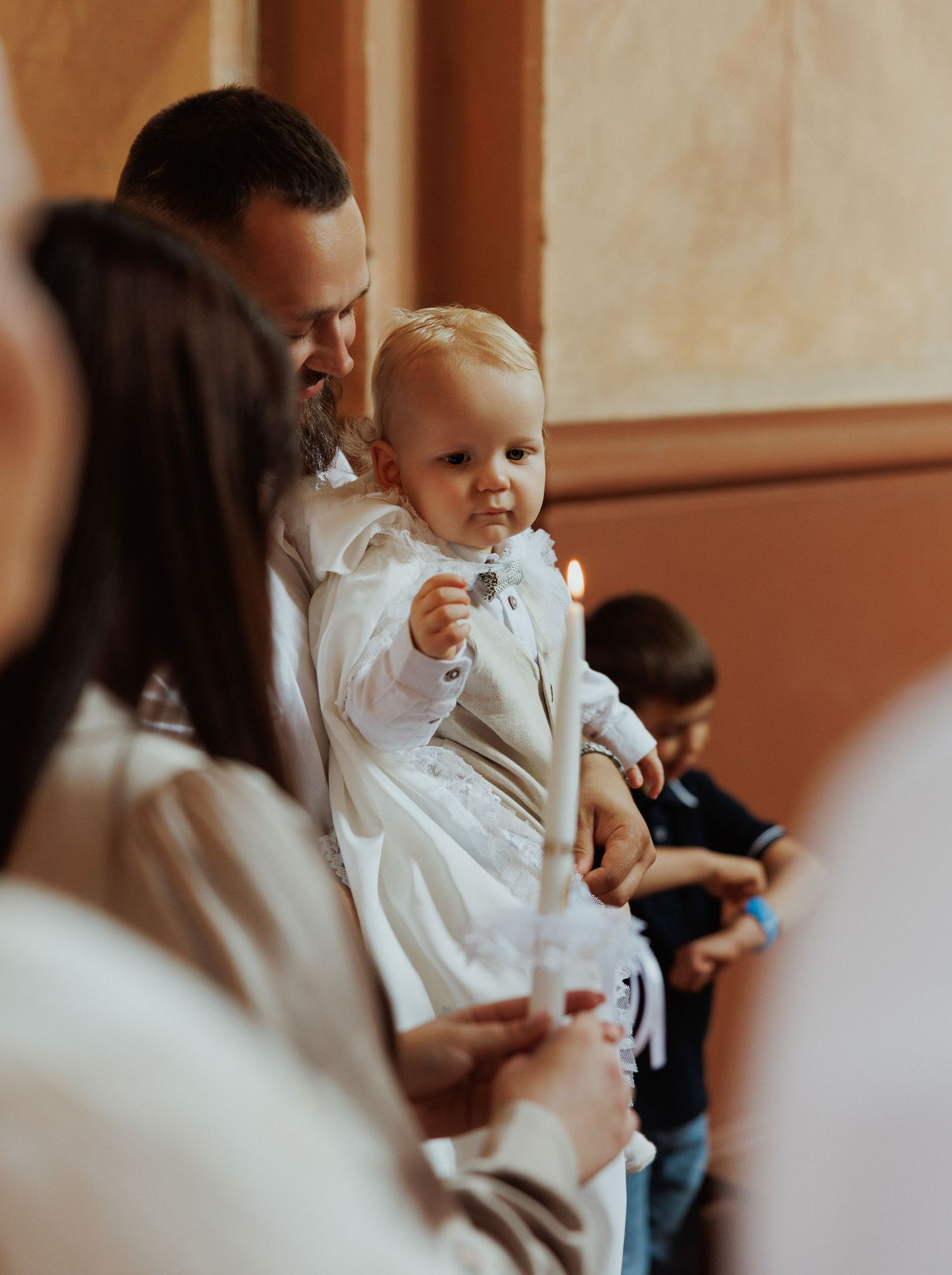 Baby in white christening gown being held during religious ceremony in warm-lit church setting.