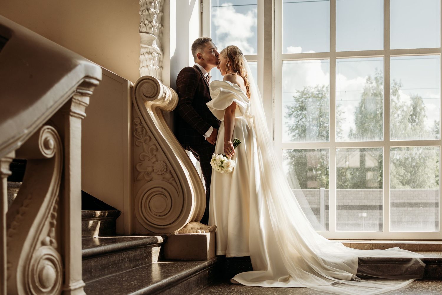 Elegant couple embraces by large windows in a bright room with ornate architectural details and flowing curtains.