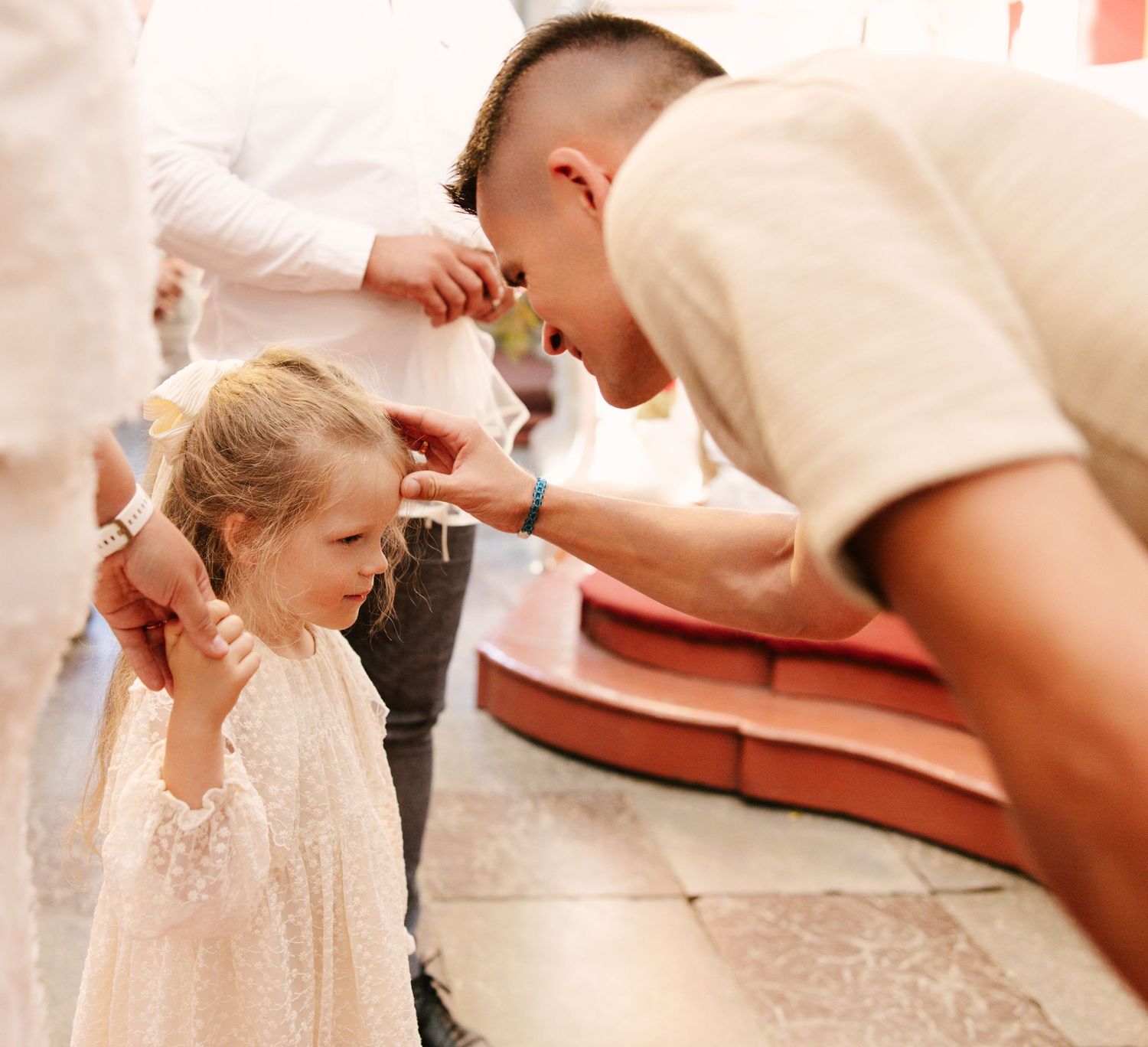 A small child in a white lace dress covers their ears while an adult leans down to speak to them.