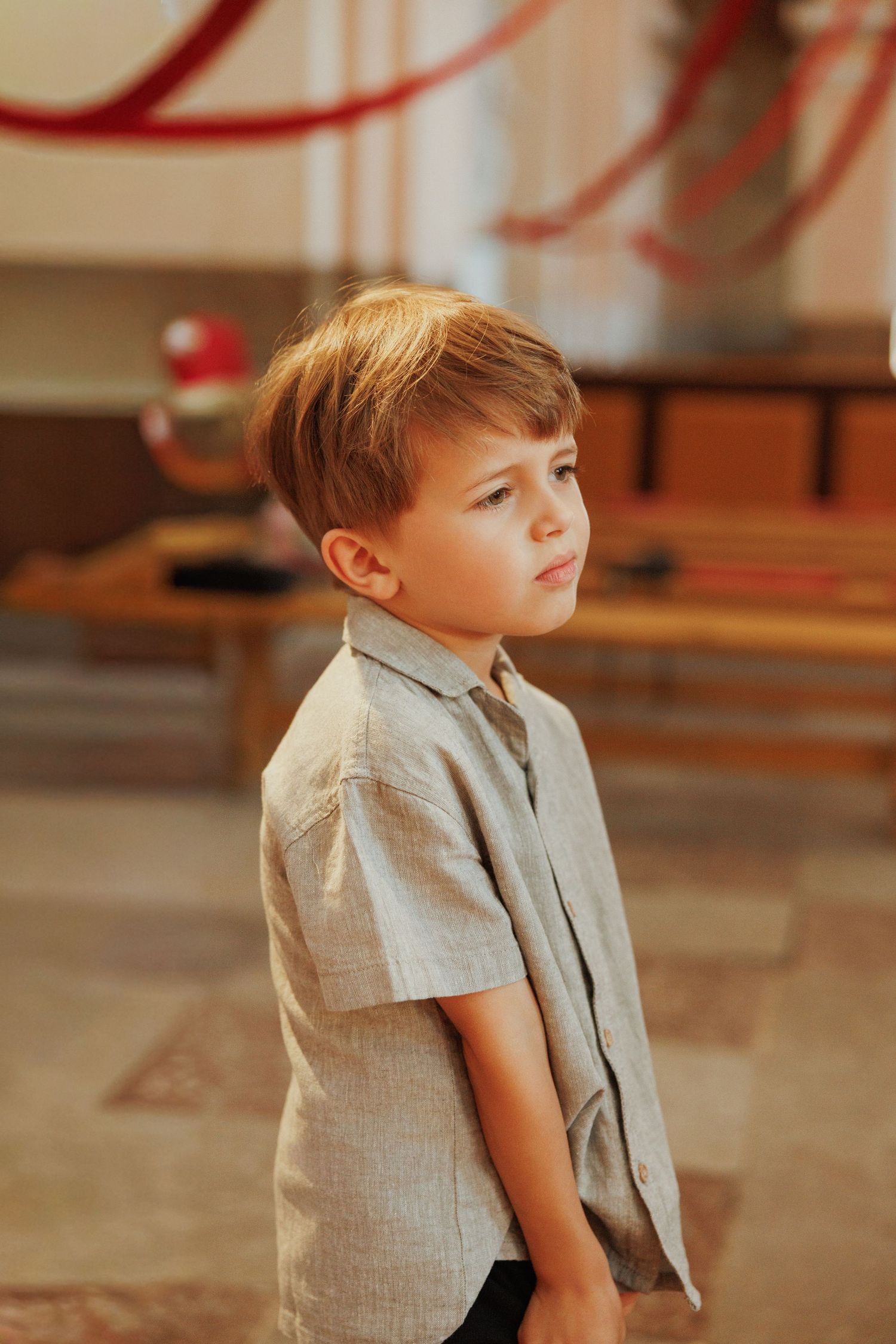 Young boy in a light gray suit jacket looks up thoughtfully against a blurred background.