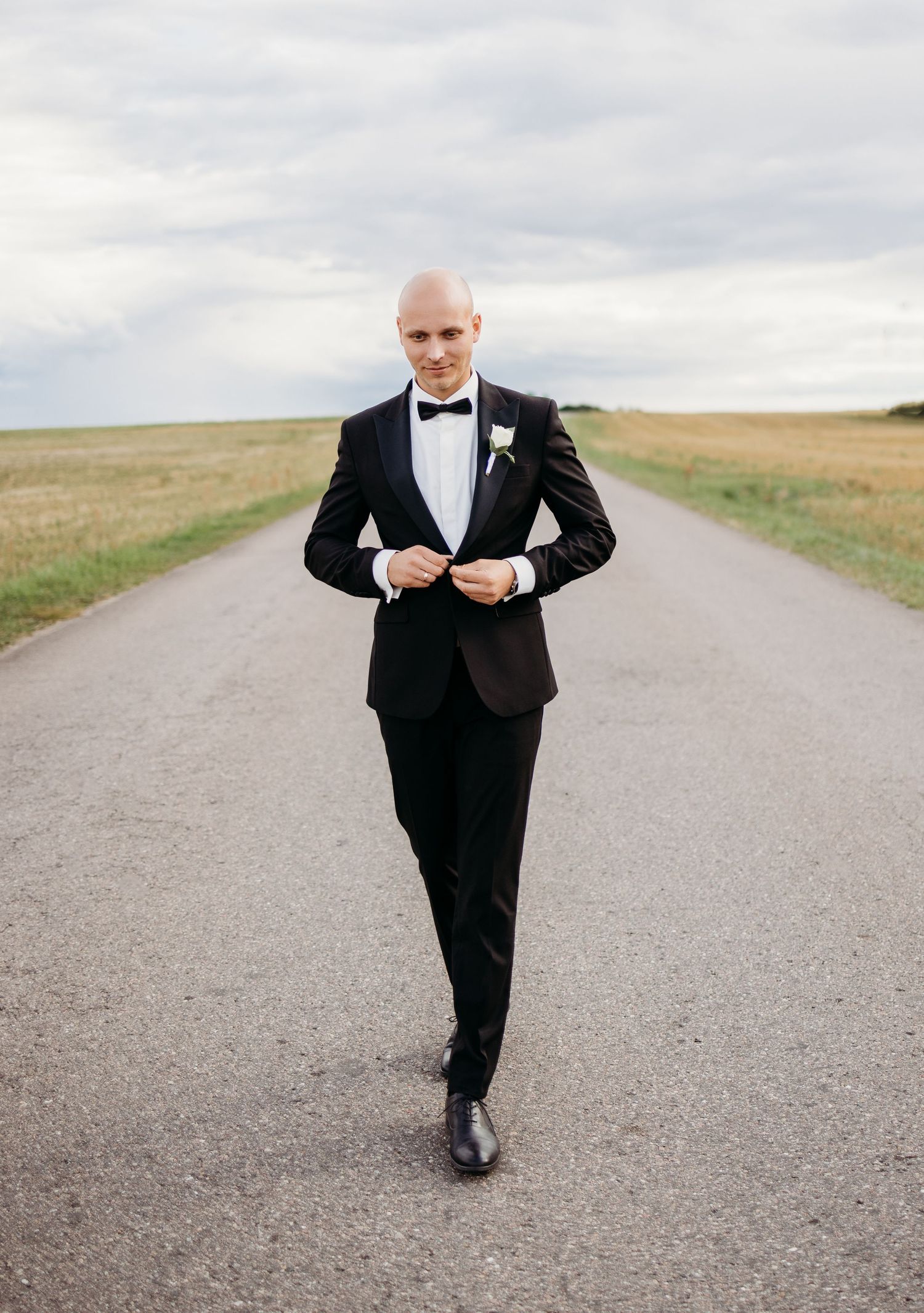 Groom in black tuxedo walks confidently down rural road during wedding photoshoot.