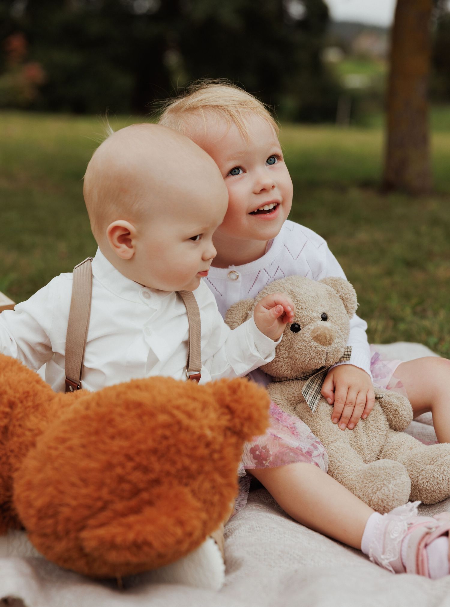 Baby lying on white blanket with teddy bears and plush toys during outdoor photography session.