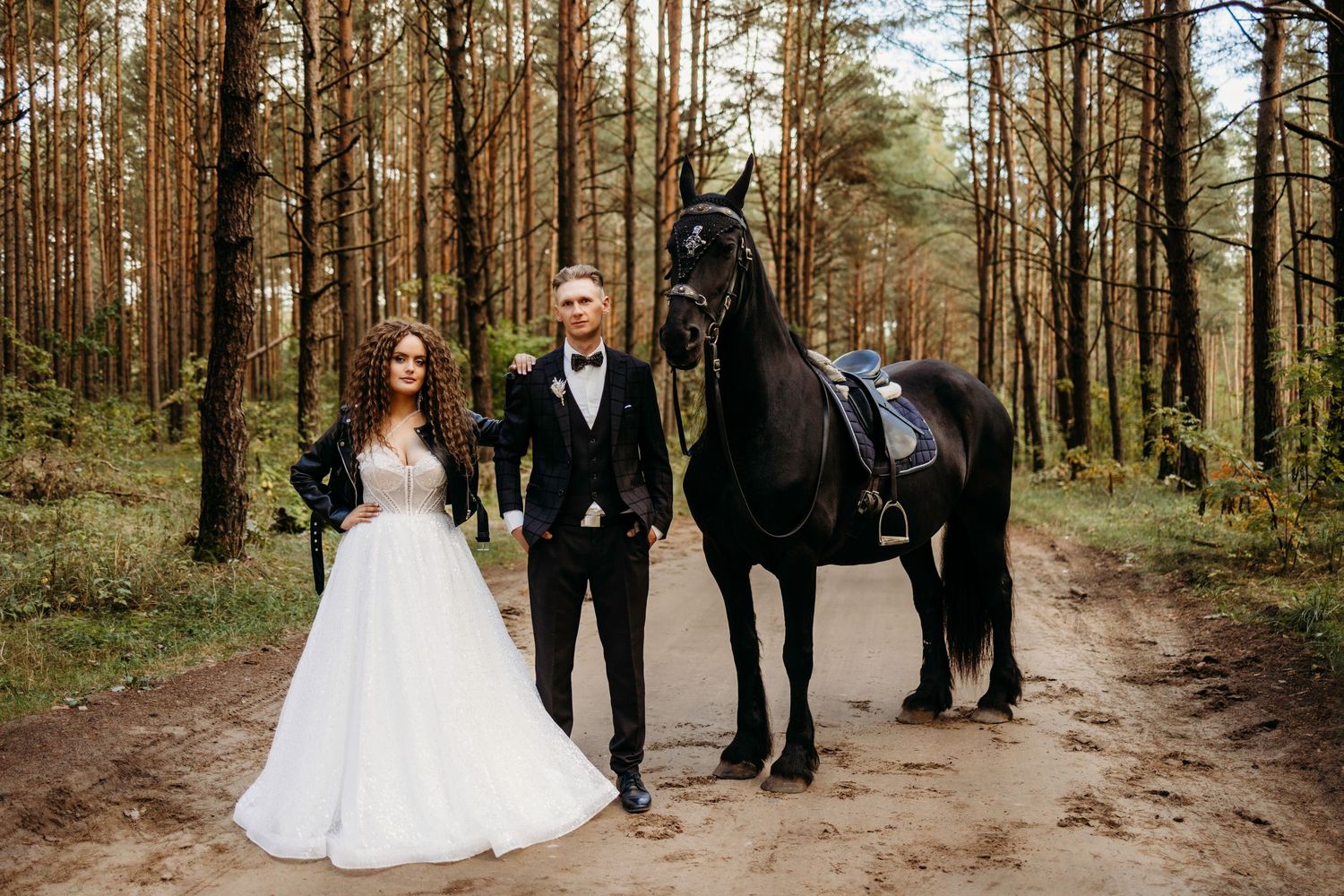 Wedding party walking through a sunlit forest path lined with tall pine trees.