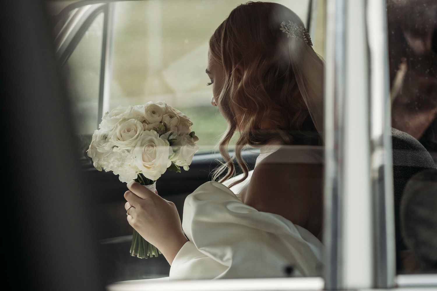 Someone in a white dress holds a bouquet of white roses while sitting in a car on their wedding day.