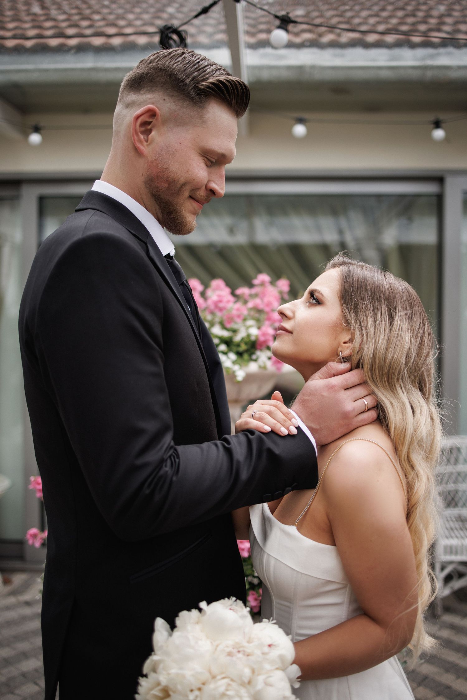 Newlyweds share tender moment in front of building with pink flowers and brick patio.