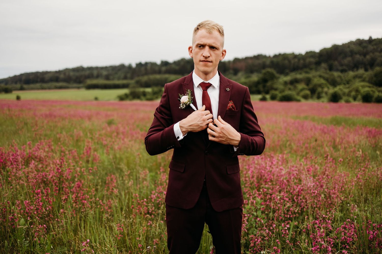 Groom in burgundy suit adjusts tie while standing in flowering meadow.