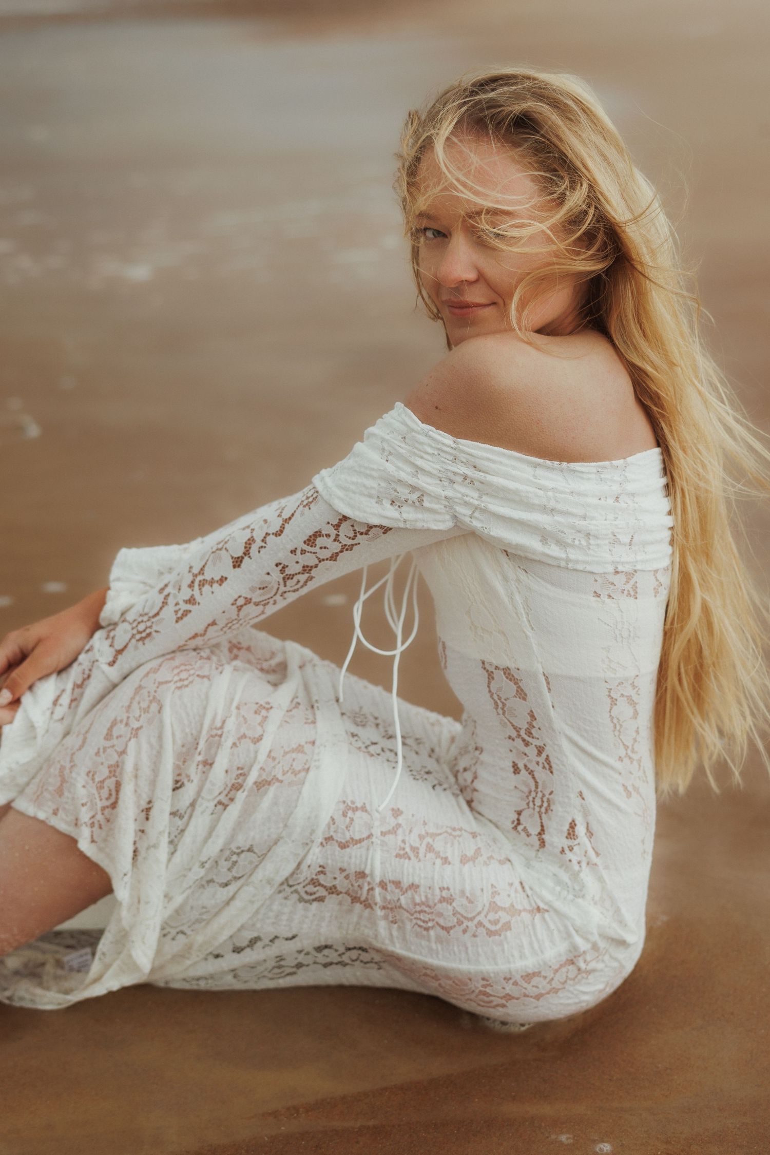 Blonde woman in white lace off-shoulder dress sitting on sandy beach shore with waves.