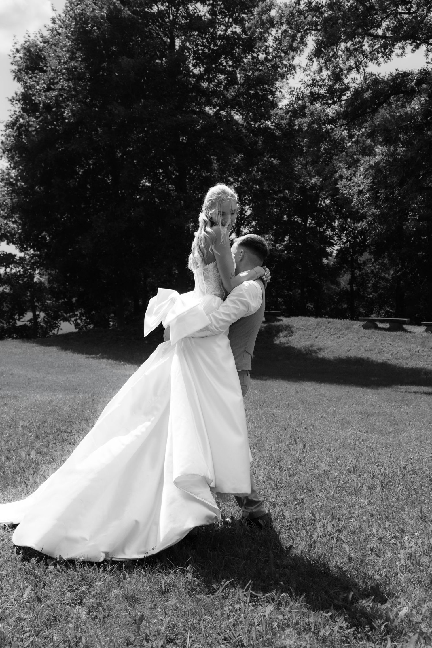 Black and white photo of groom lifting bride in white wedding gown outdoors on green grass with trees in background.