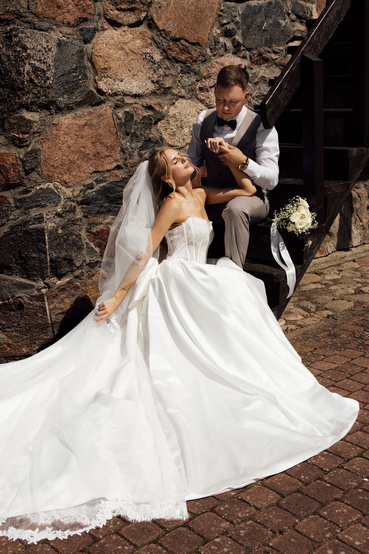 Bride in white ball gown sits on stone steps as groom in gray vest holds her hand at rustic venue.