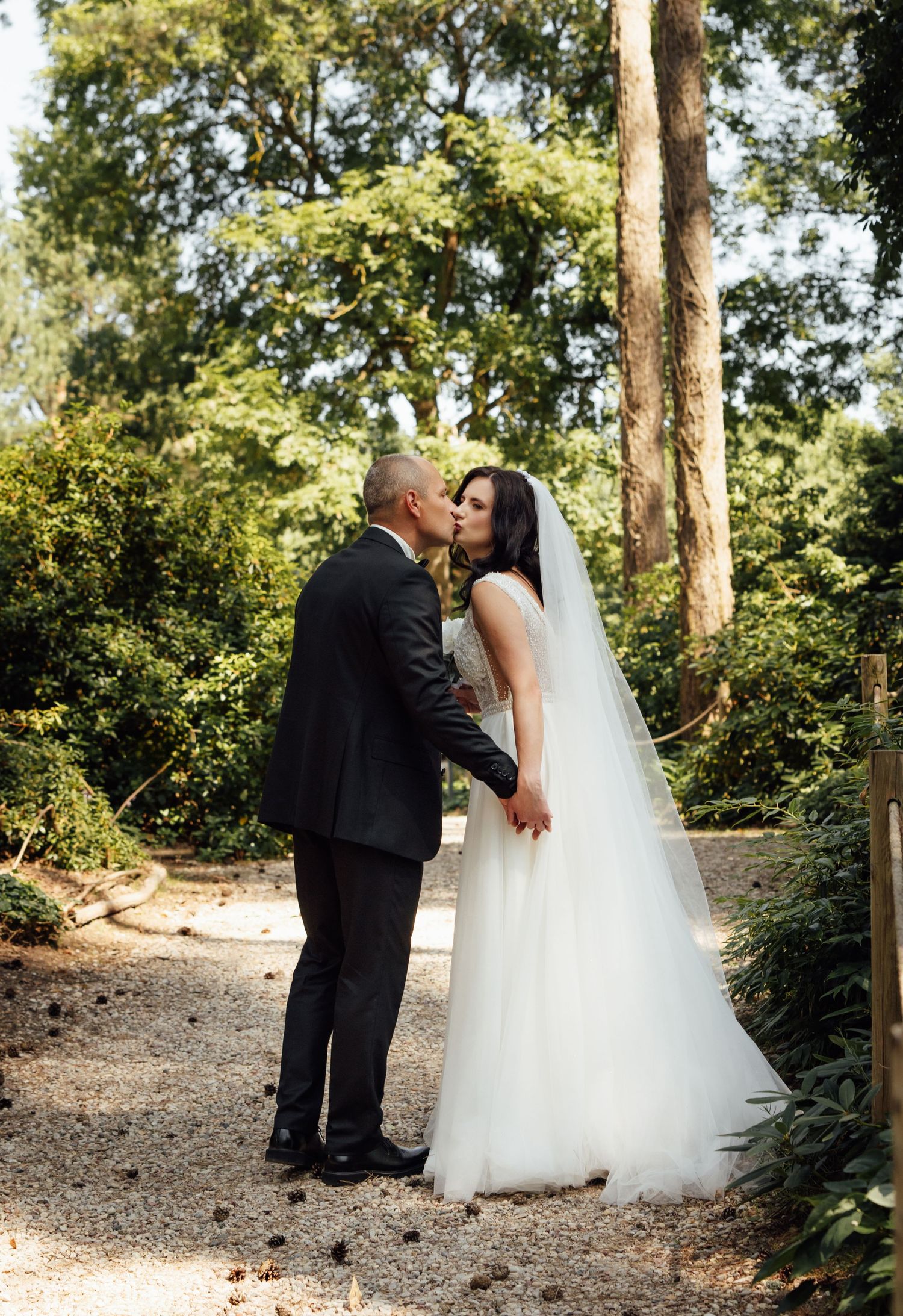 Bride and groom sharing a kiss on a garden path surrounded by lush green trees on their wedding day.