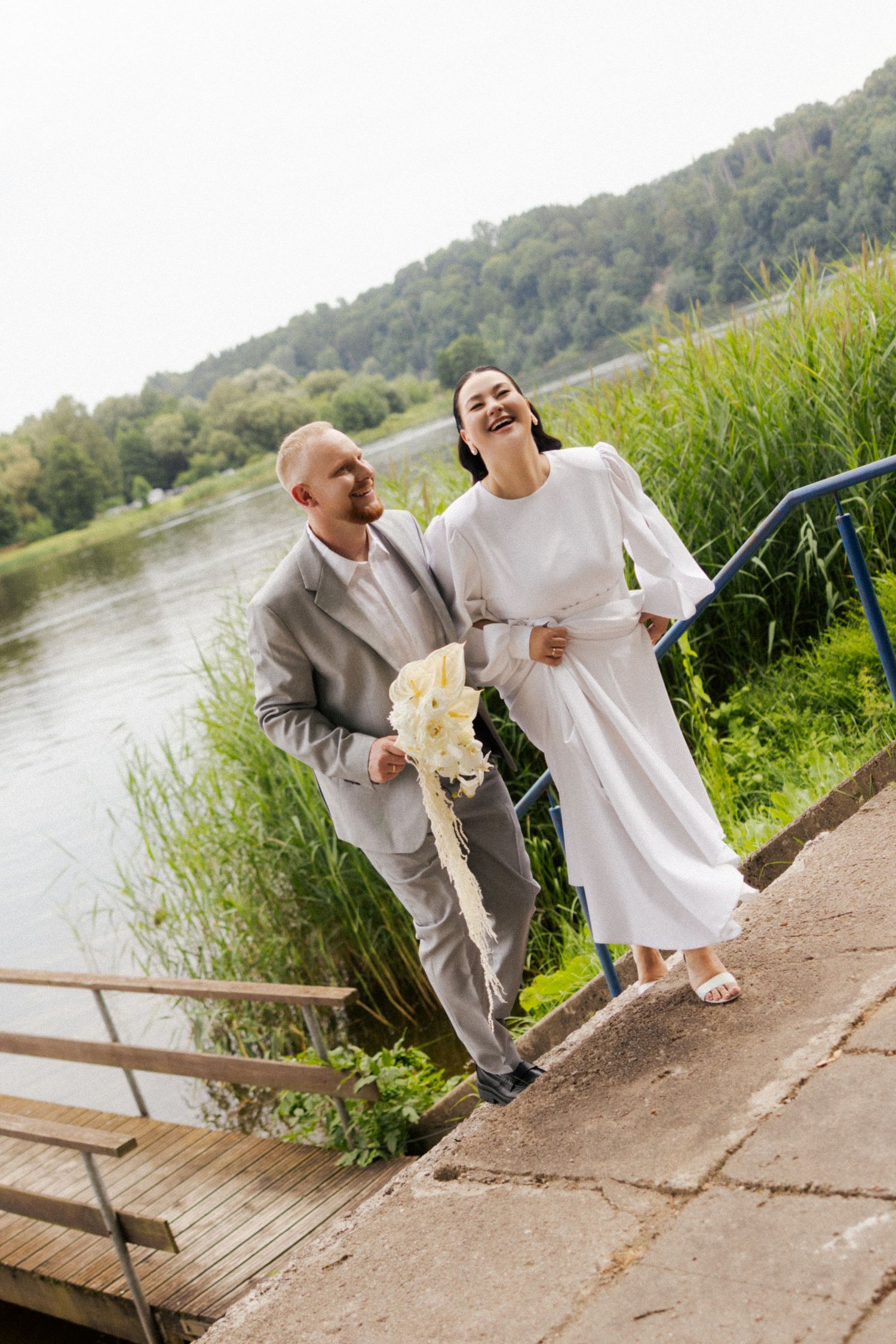 Happy newlywed couple walking by a lakeside, bride in white dress holding bouquet, groom in gray suit, lush greenery background.
