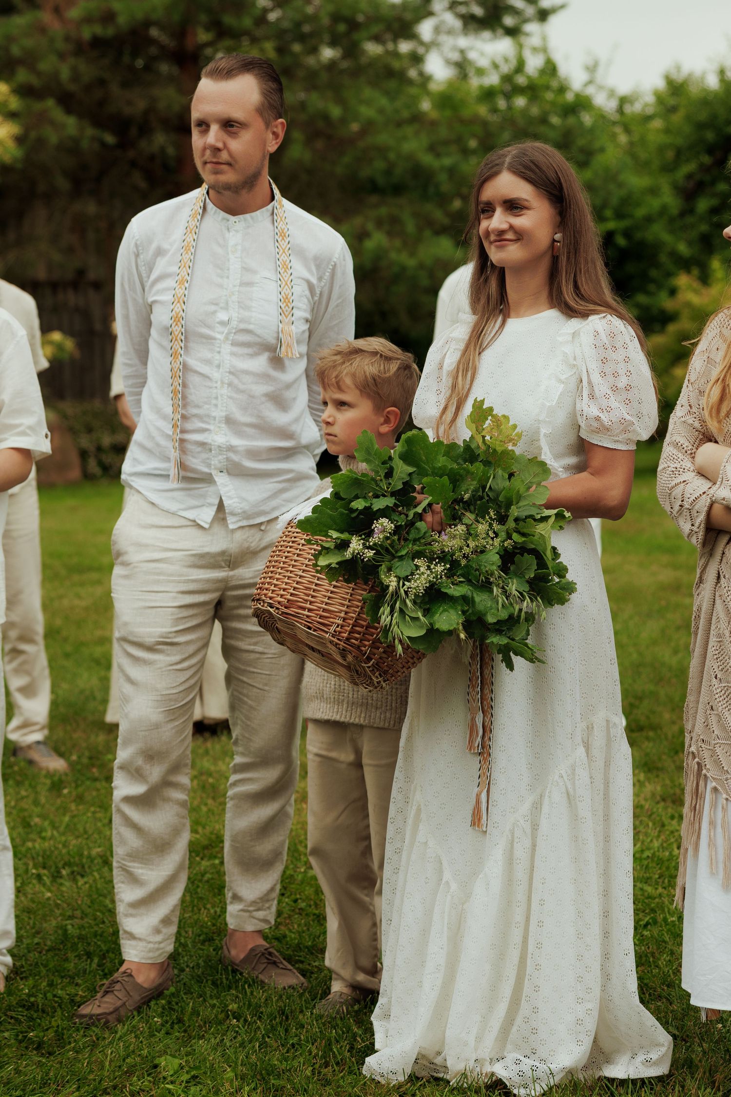 Couple in white attire with a boy holding a wicker basket of greenery at an outdoor wedding ceremony.